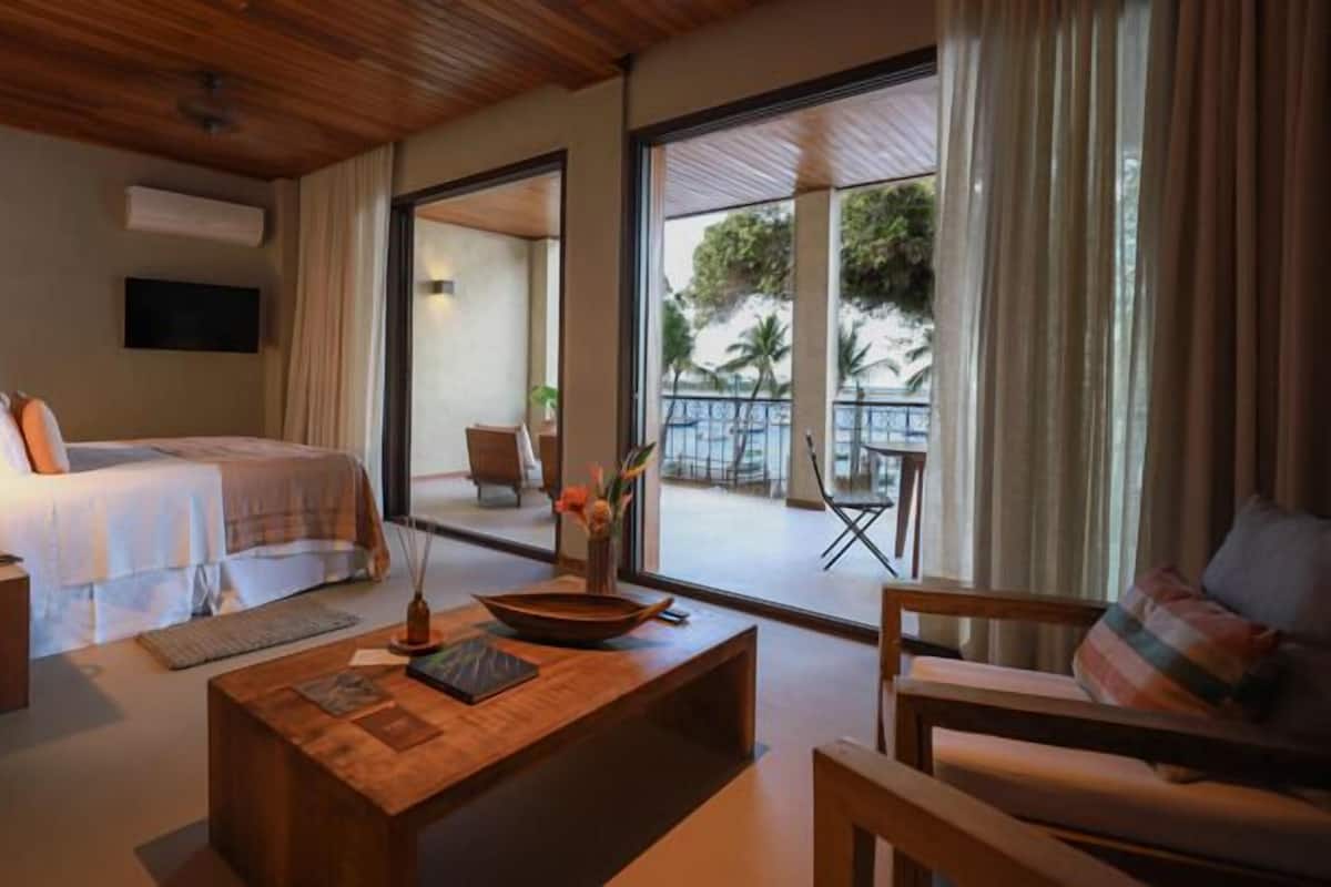 Een hotelkamer met een bed, een houten bijzettafeltje, een bank en een balkon met uitzicht op palmbomen.
