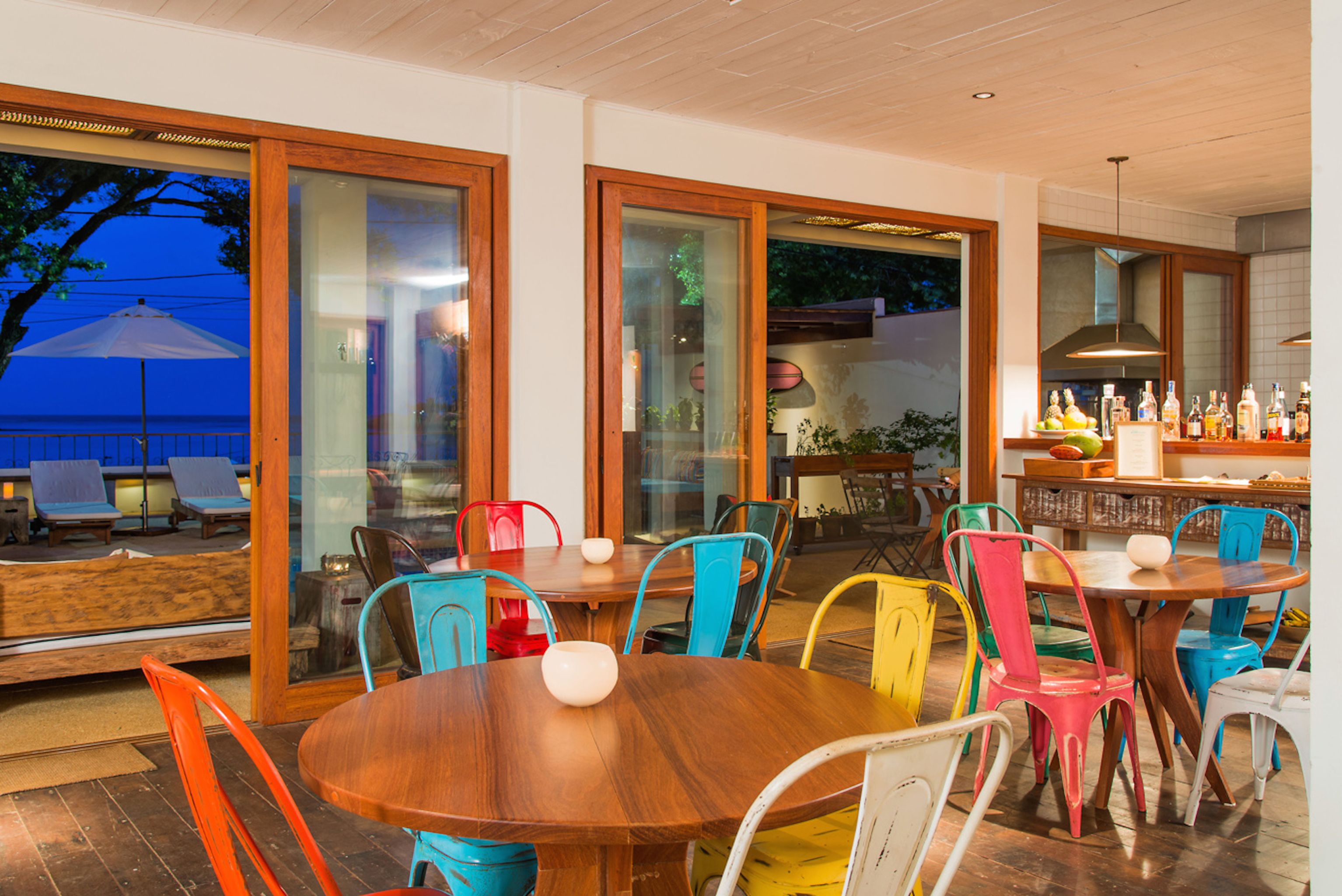 Zona pranzo con tavoli in legno e sedie colorate, ampie finestre con porte in vetro e un'area bar con bottiglie.