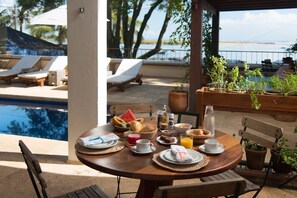 A dining table with breakfast items, a pool, and lounge chairs in the background. - OITI by Barracuda (Itacaré)