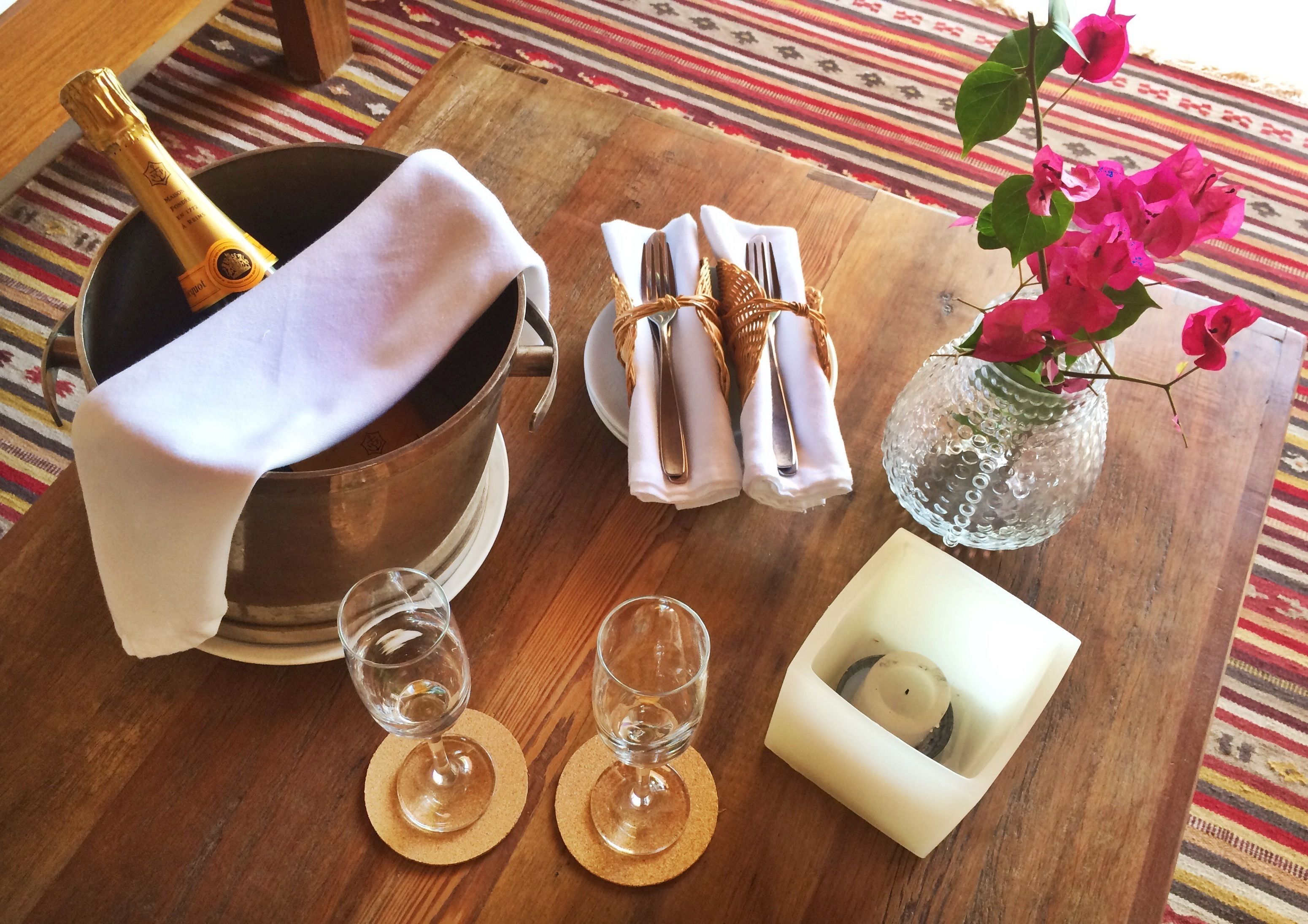A wooden table with a champagne bottle, two wine glasses, a vase with pink flowers, and a candle holder.