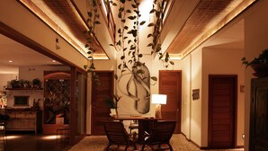 Vestíbulo de hotel con zona de comedor, mesa y sillas, un cuadro grande en la pared y plantas colgantes.