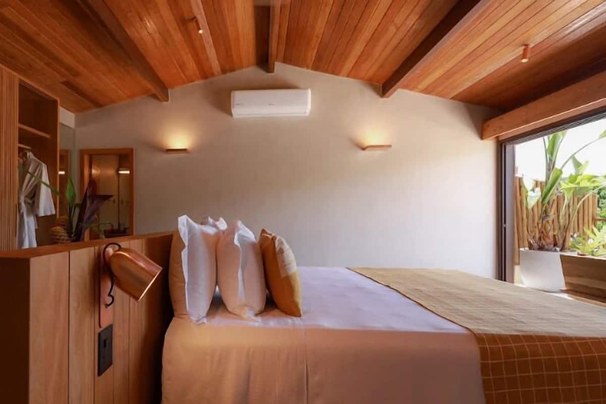 Een slaapkamer met een houten plafond, een groot bed met kussens, een nachtkastje en een raam met planten.