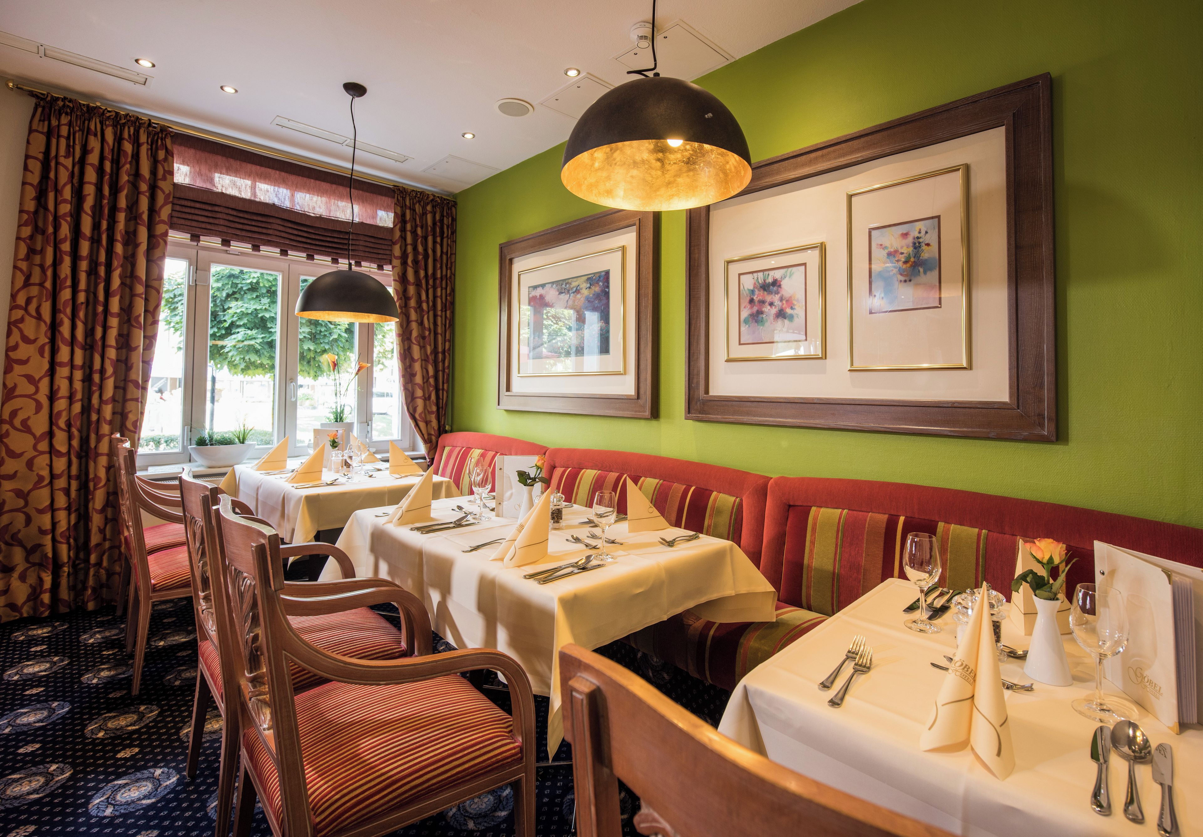 Interior de restaurante con mesas dispuestas para comer, una pared verde con cuadros enmarcados y una alfombra estampada.