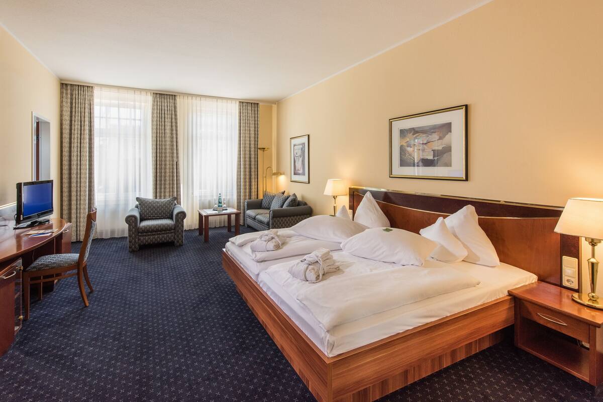 Ein Hotelzimmer mit einem großen Bett, einem Schreibtisch mit Computer, einem Stuhl, einem kleinen Tisch und zwei Lampen.