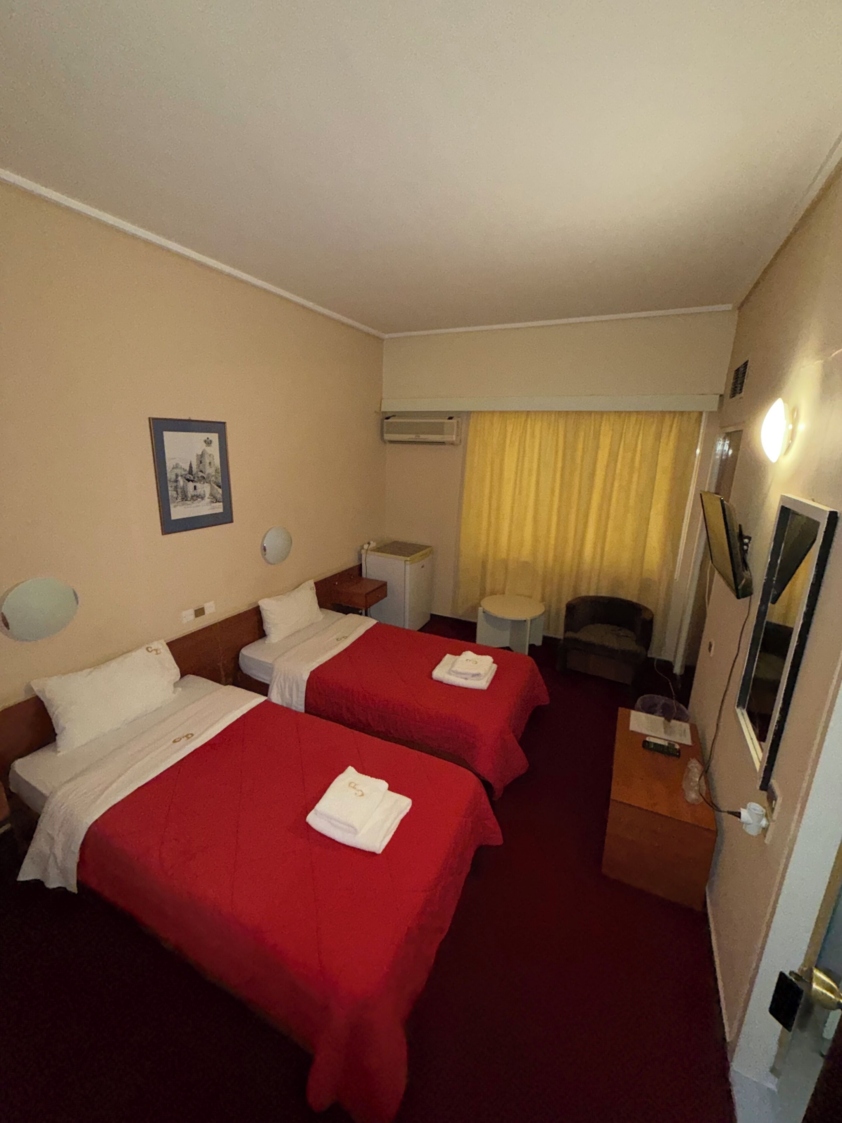 Quarto de hotel com duas camas, uma mesa pequena, uma cadeira, ar condicionado, um quadro na parede e uma janela com cortinas.