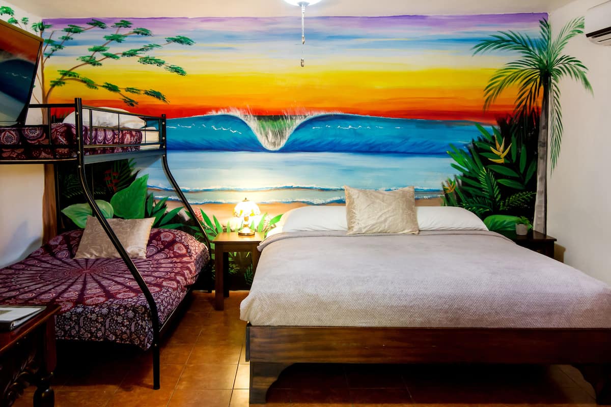 Une chambre avec un lit superposé, un lit simple et une fresque murale représentant un coucher de soleil sur l’océan.