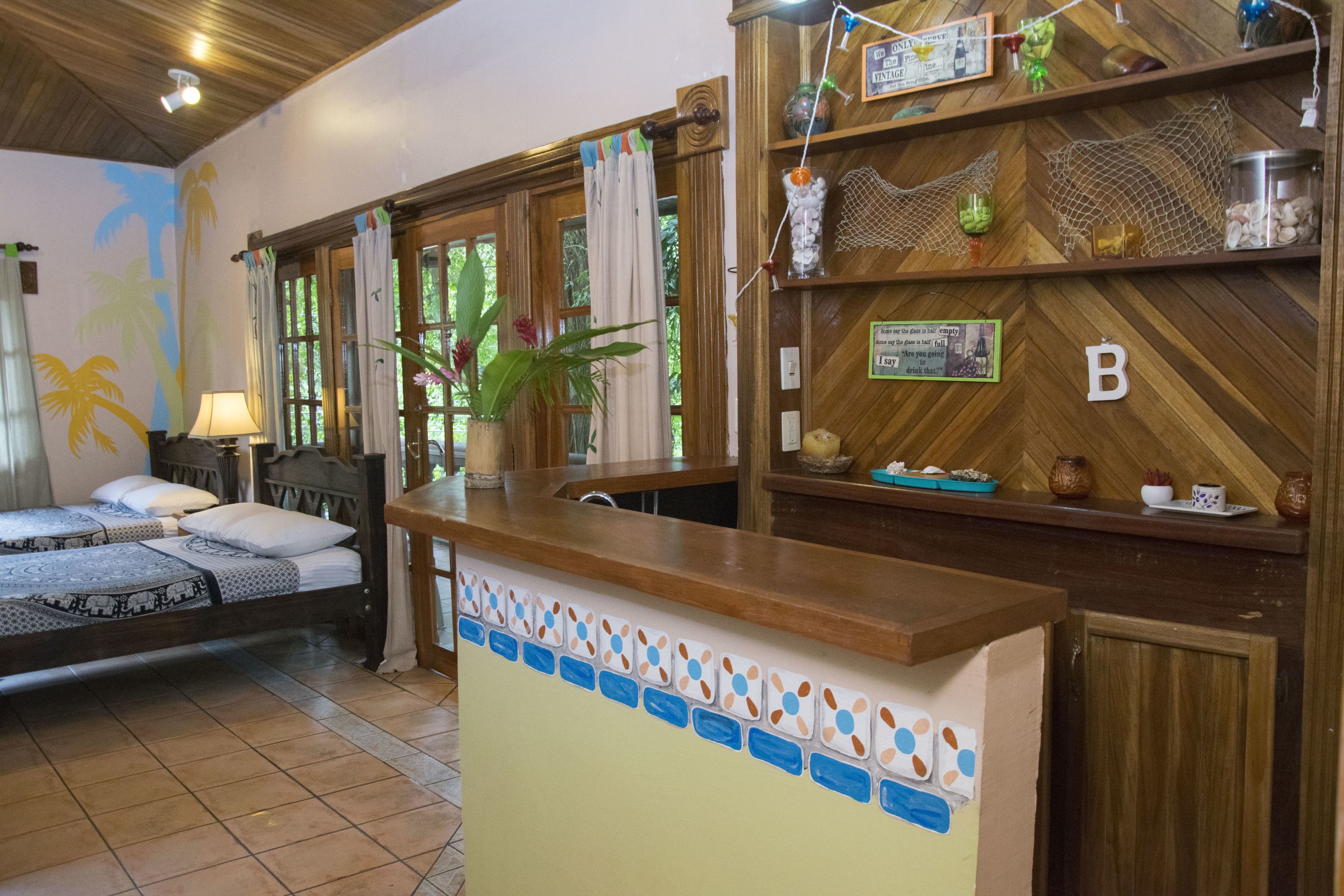 A hotel room with a bed, a desk, a window with curtains, a plant, and a wooden wall with shelves.