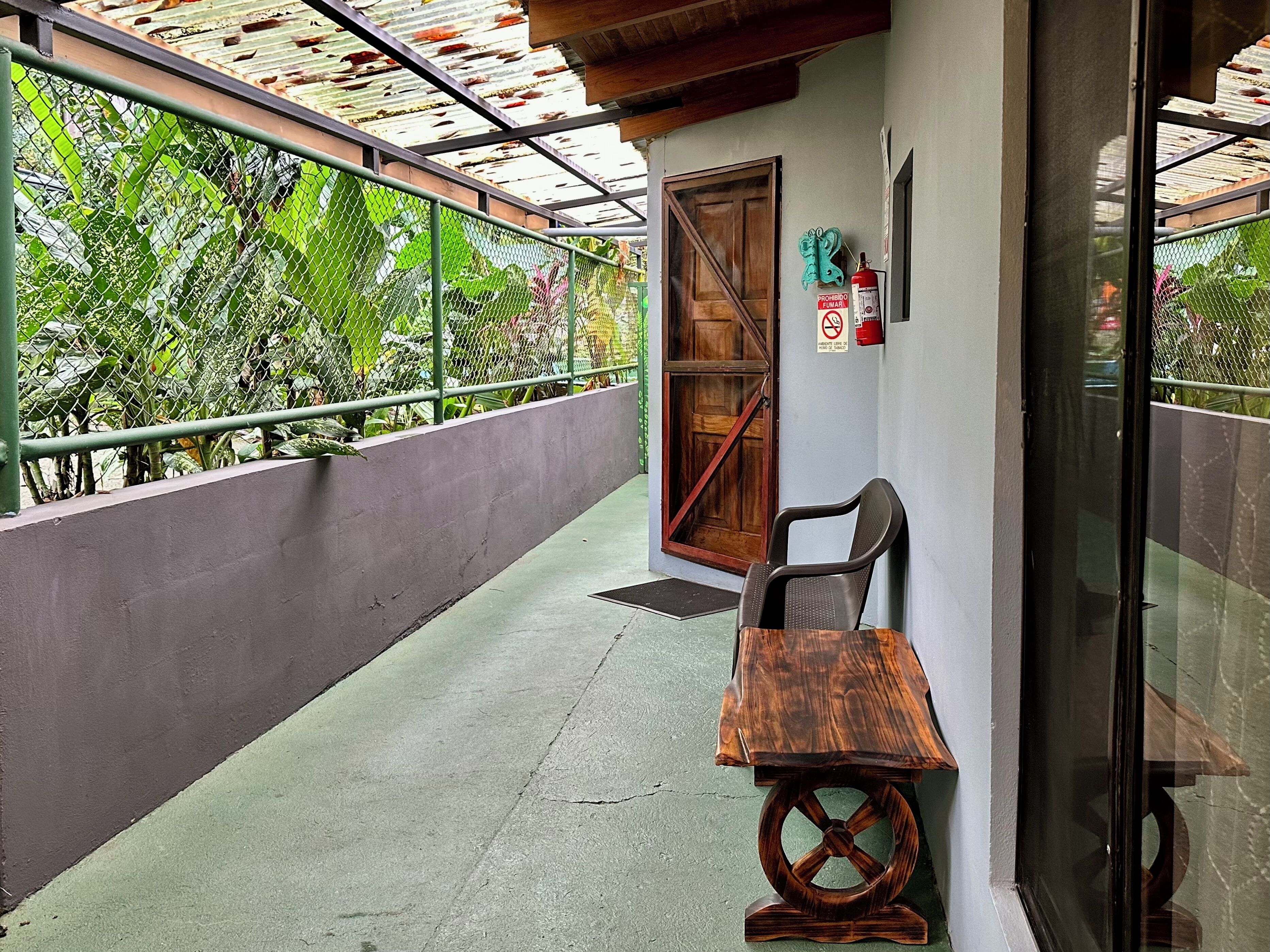 A balcony with a wooden table and chair, a metal railing, and a view of lush greenery.
