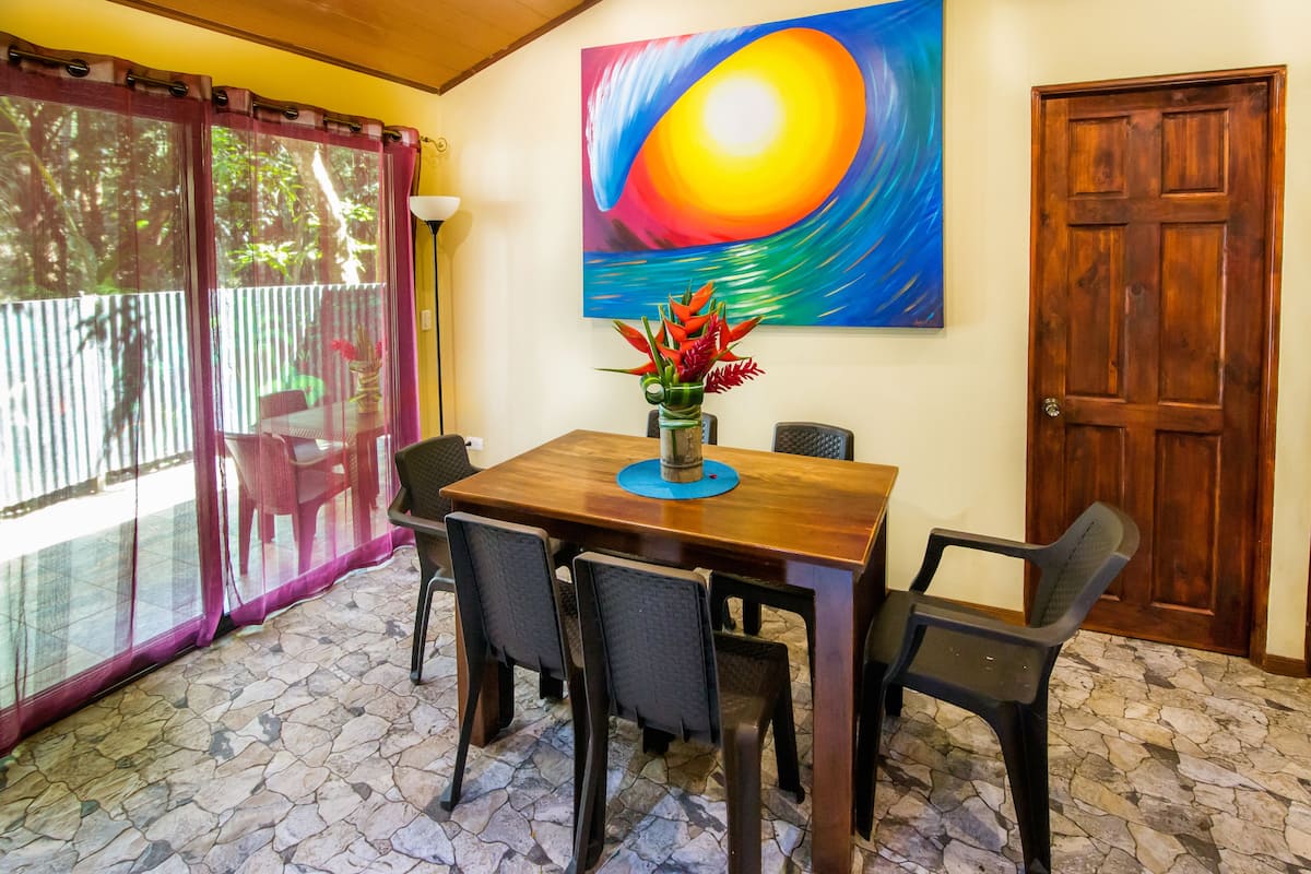 Un coin repas avec une table en bois, quatre chaises, un vase de fleurs et un tableau représentant un coucher de soleil accroché au mur.