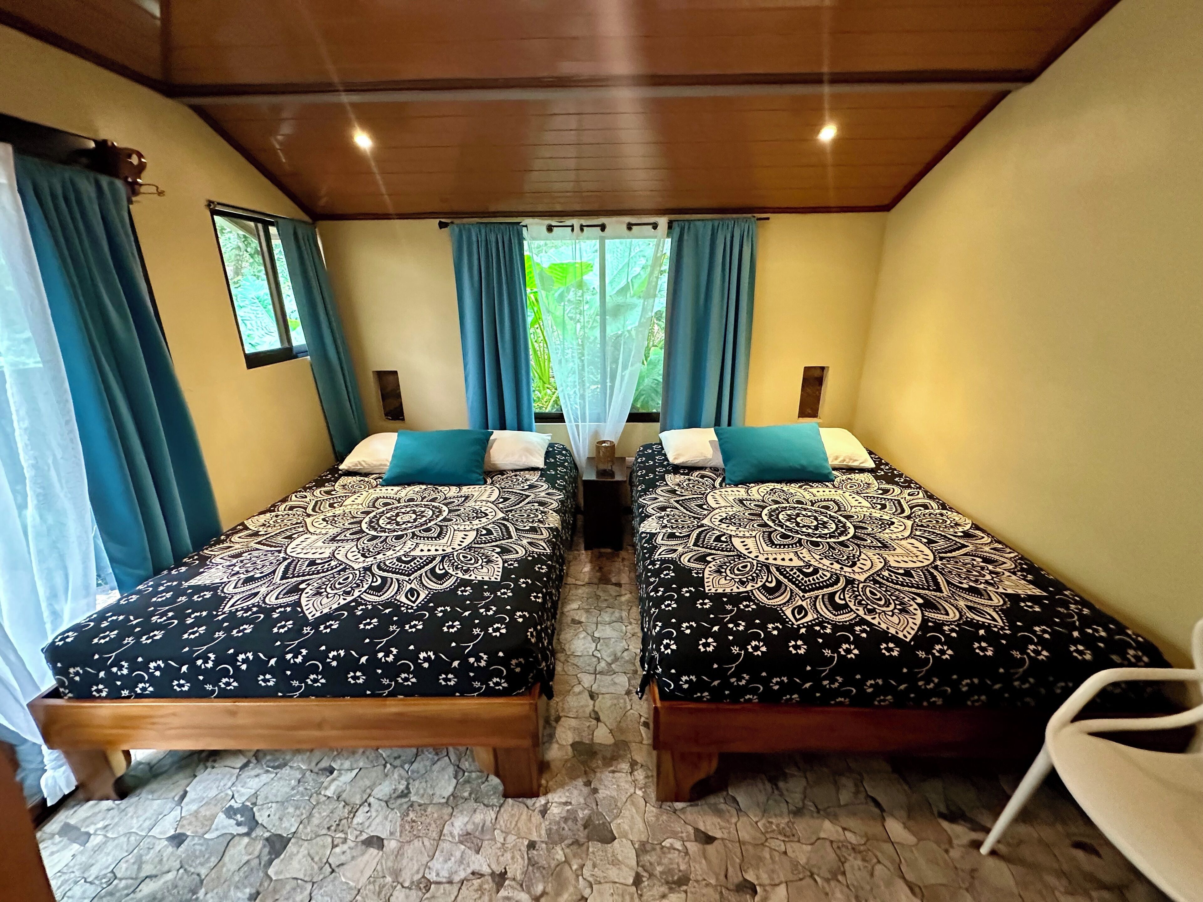 A room with two beds, each with patterned bedding, a stone floor, wooden ceiling beams, and a window with curtains.