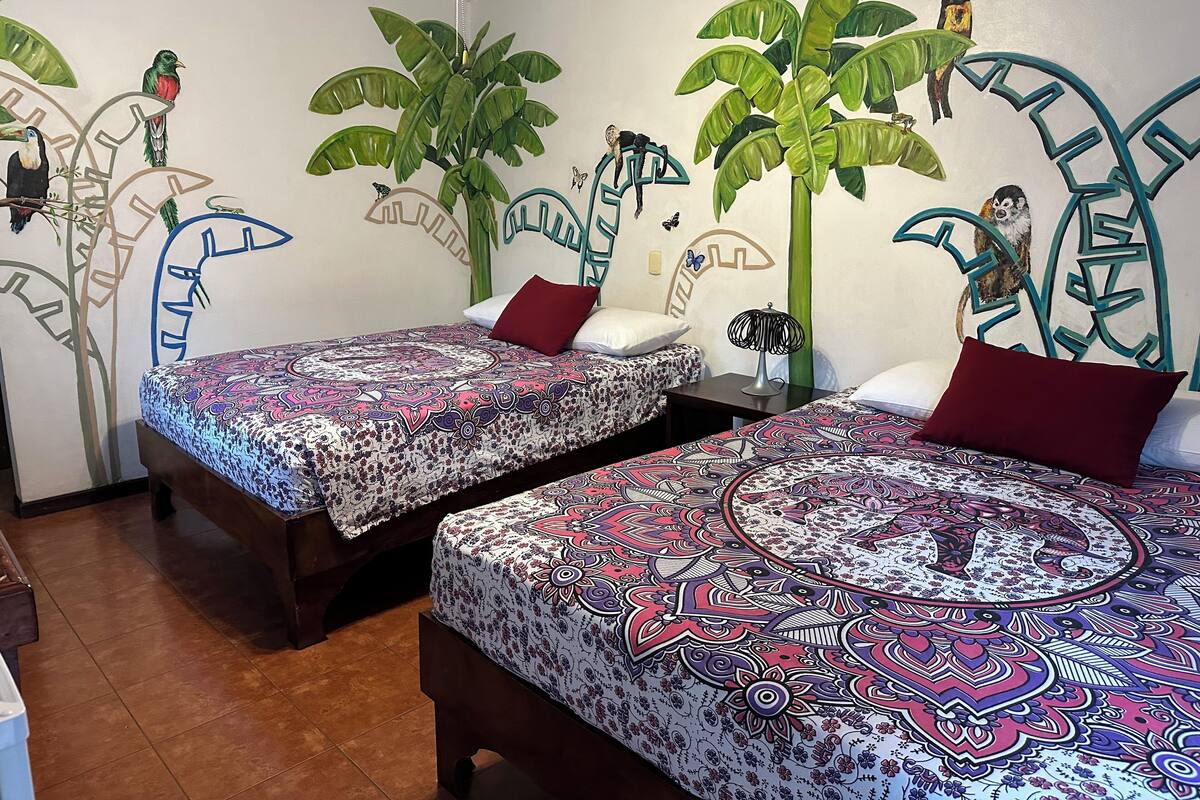 Une chambre avec deux lits, chacun agrémenté de draps à motifs, un mur décoré d’une fresque représentant des oiseaux et des plantes, et un luminaire au plafond.