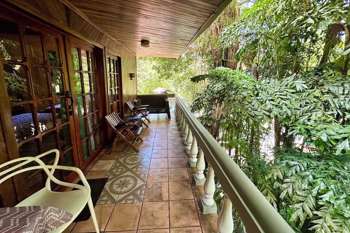 Un espace extérieur aménagé, abrité et doté de mobilier en bois et d’un sol carrelé, entouré d’une végétation luxuriante.