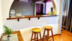 Un coin bar avec un comptoir en bois, deux tabourets, une plante en pot et une petite table sur laquelle se trouve une télécommande.