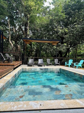 Una piscina rectangular con agua azul cristalina, rodeada de un deck de madera y sillas, en un entorno verde y exuberante.