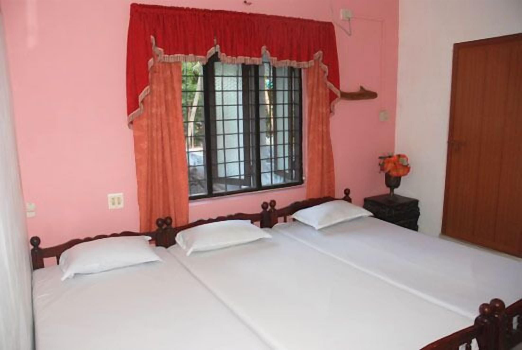 A room with two beds, a window with curtains, a nightstand with a lamp, and a wooden door.