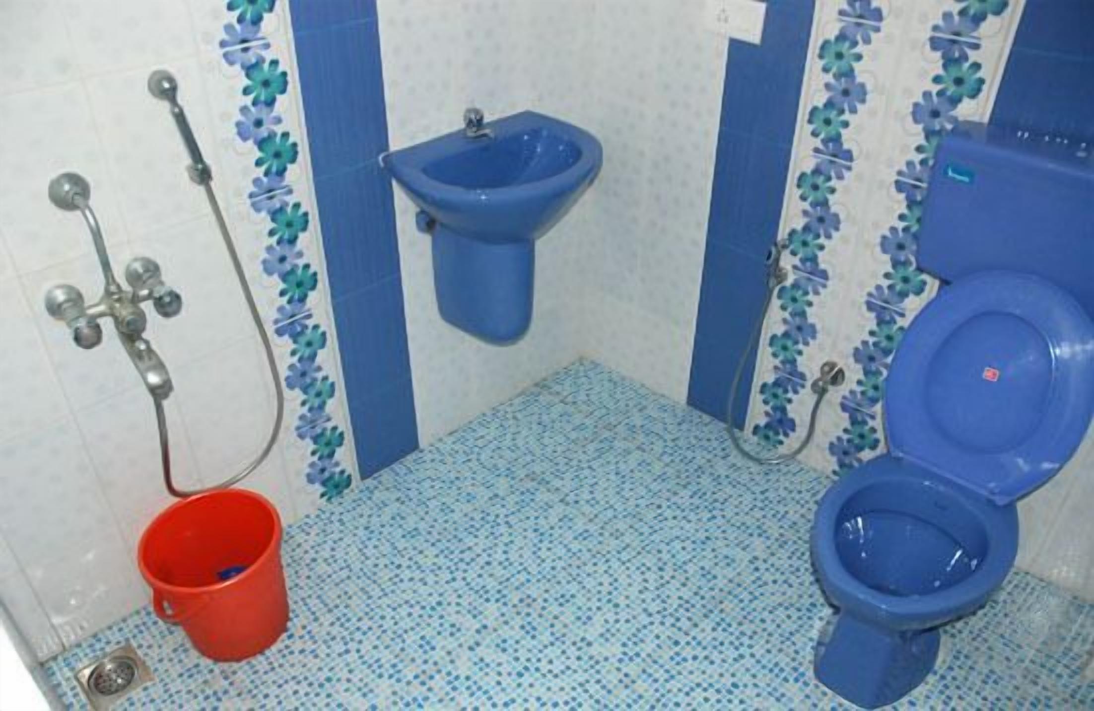 Une salle de bain avec des toilettes bleues, un bidet bleu et un seau rouge posé sur le sol.