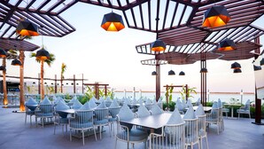 Un área de comedor al aire libre con mesas y sillas, una estructura de pérgola y lámparas colgantes.