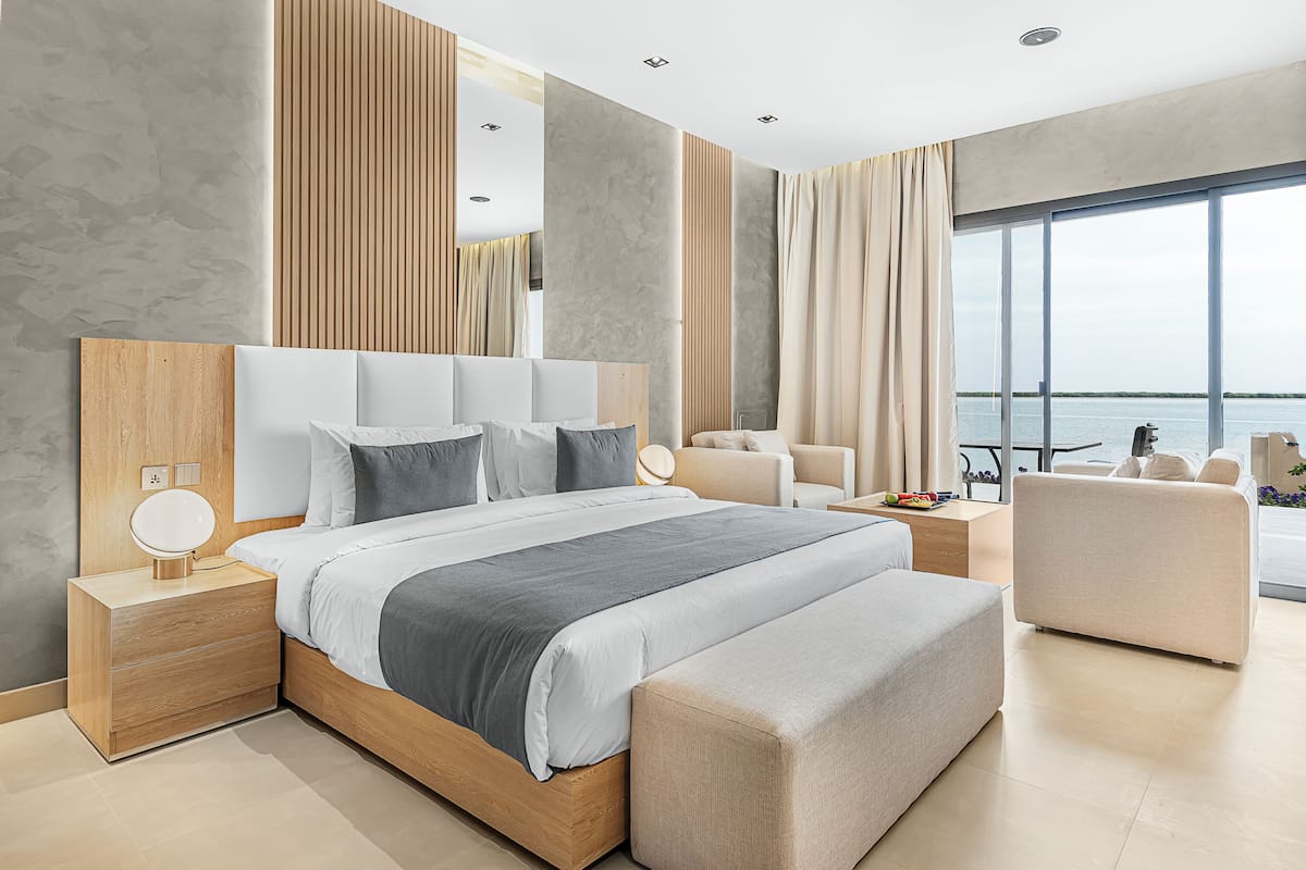 A modern bedroom with a large bed, bedside tables, a seating area, and a view of the water.