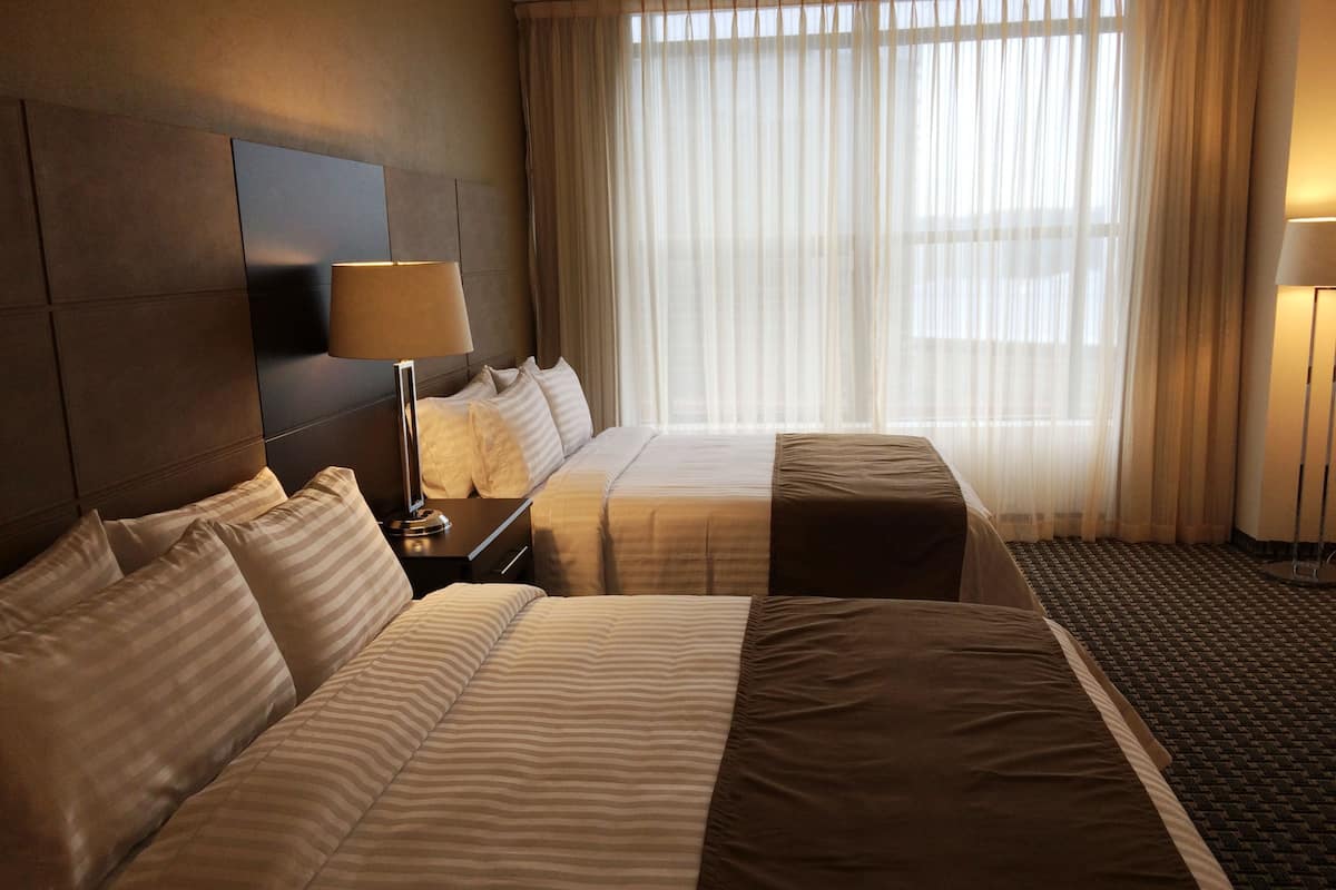 A hotel room with two beds, a nightstand, a lamp, and a large window with sheer curtains.