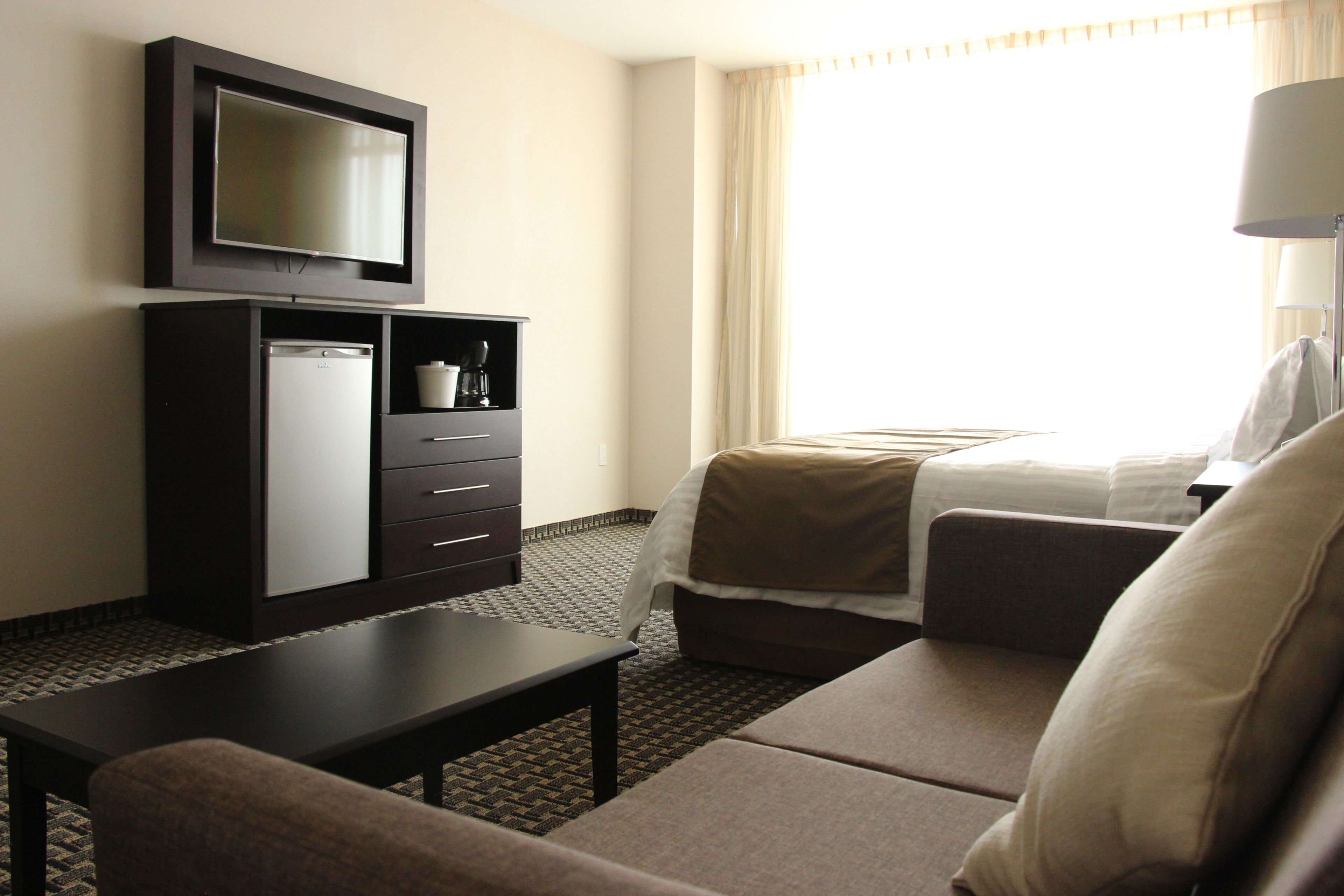 A hotel room with a television, a minibar, a bed with a white comforter, a sofa, and a table.