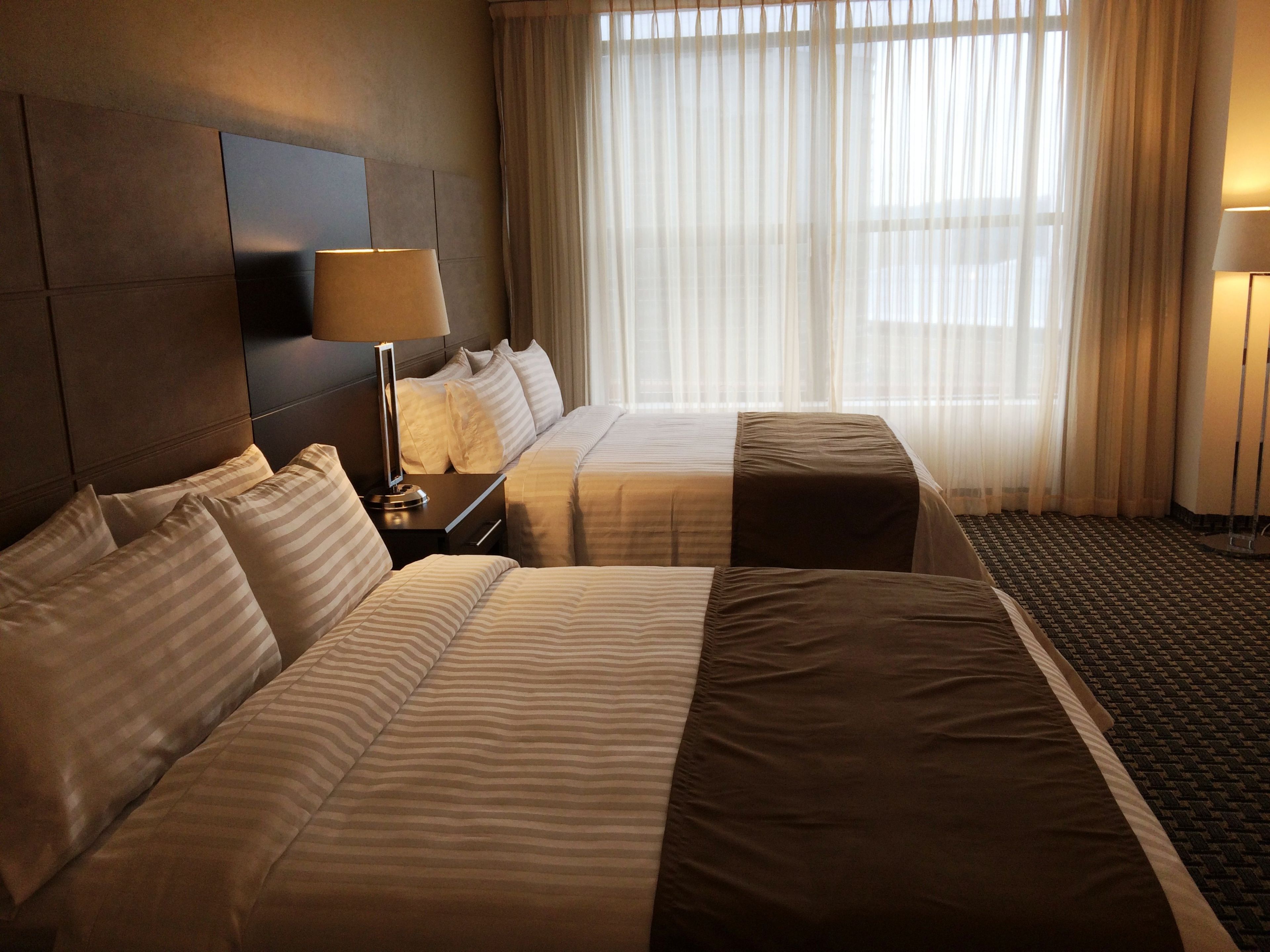 Ein Hotelzimmer mit zwei Betten, einem Nachttisch mit Lampe, einem großen Fenster mit durchsichtigen Vorhängen und einem Teppichboden.