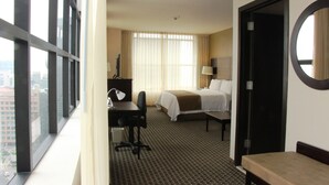 Ein Hotelzimmer mit Bett, Schreibtisch, Stuhl und Blick auf die Stadt durch große Fenster.