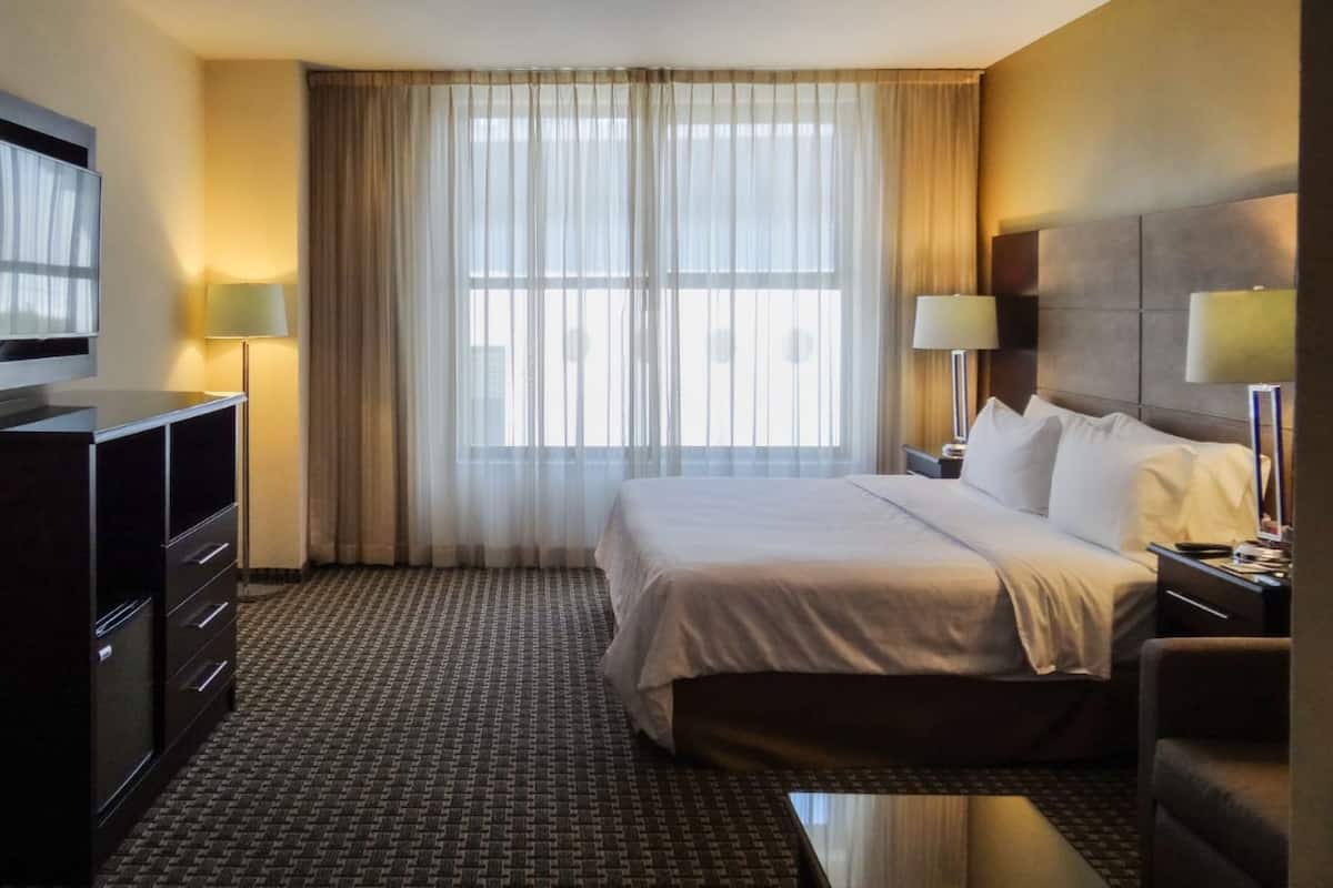 A hotel room with a large bed, a TV mounted on the wall, a desk with a lamp, and a window with sheer curtains.