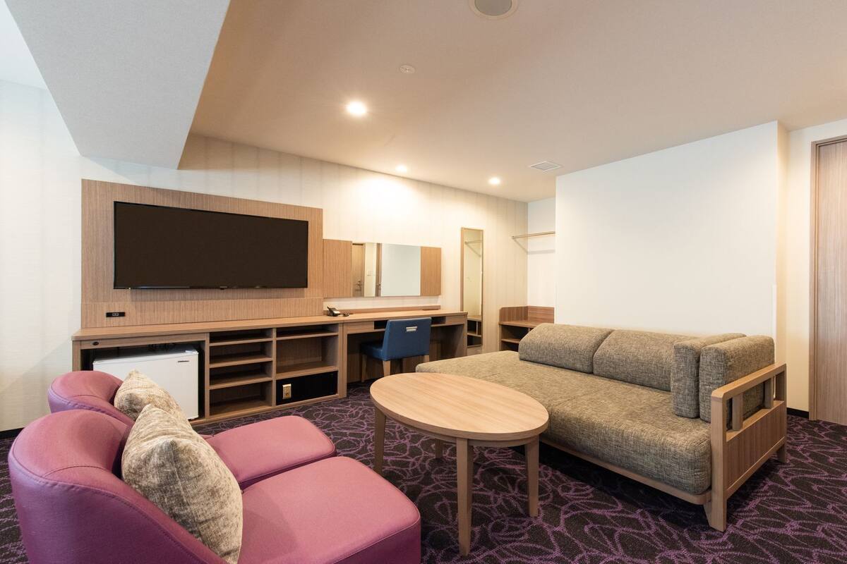 A hotel room with a flat-screen TV, a sofa, a small table, a desk, a chair, and a wardrobe.
