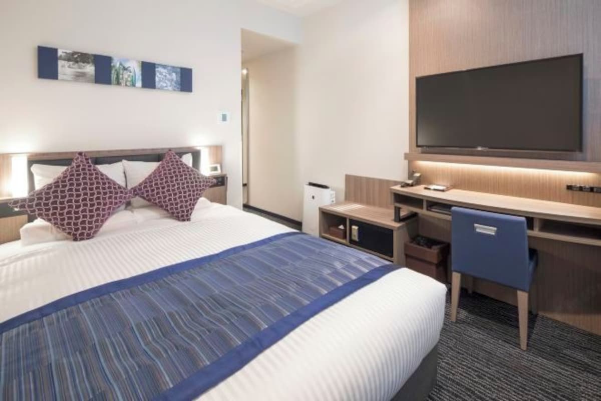 A hotel room with a large bed, a desk with a chair, a flat-screen TV, and a small desk lamp.