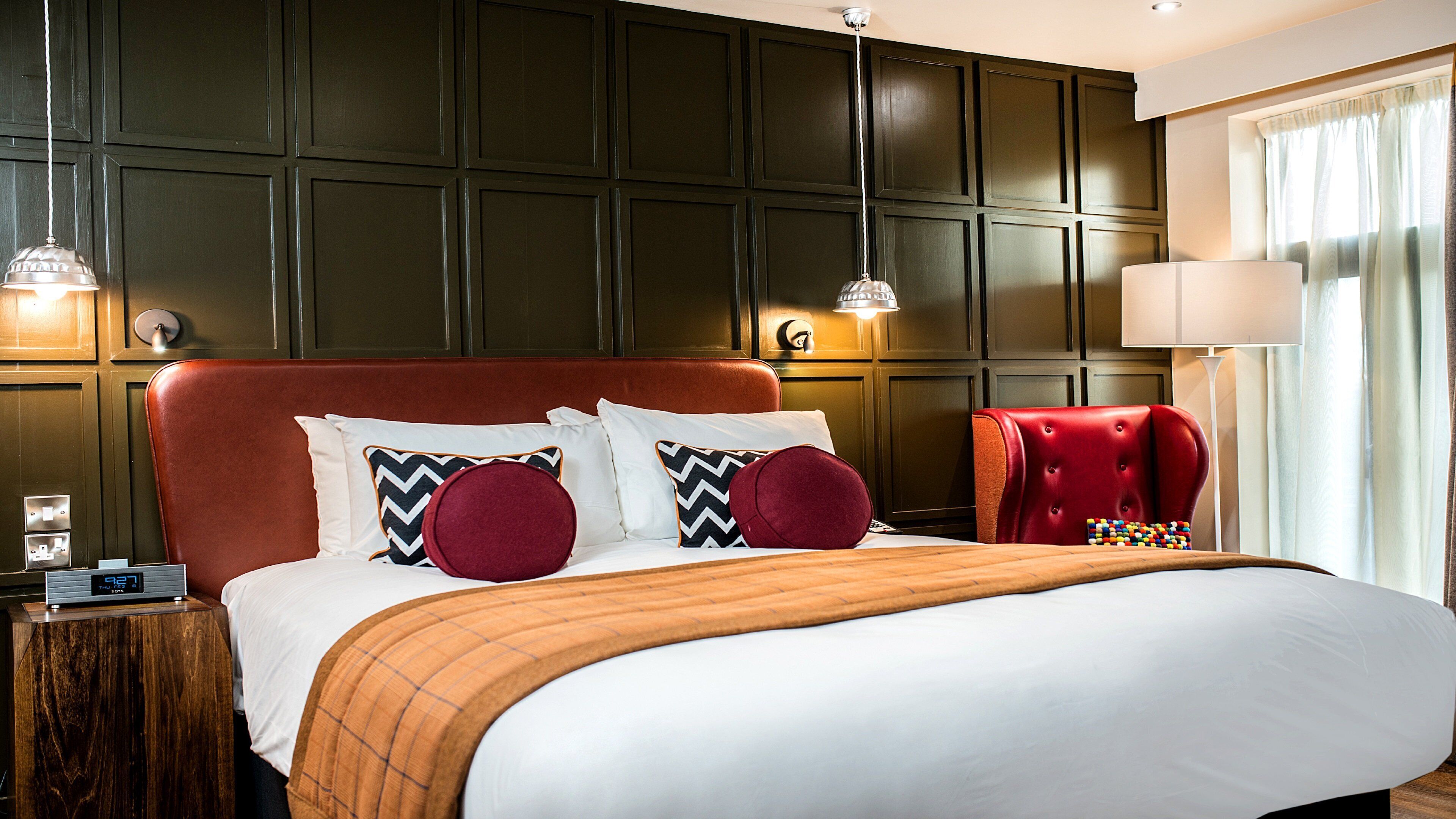 A hotel room with a large bed, a red upholstered chair, and a nightstand with a clock.