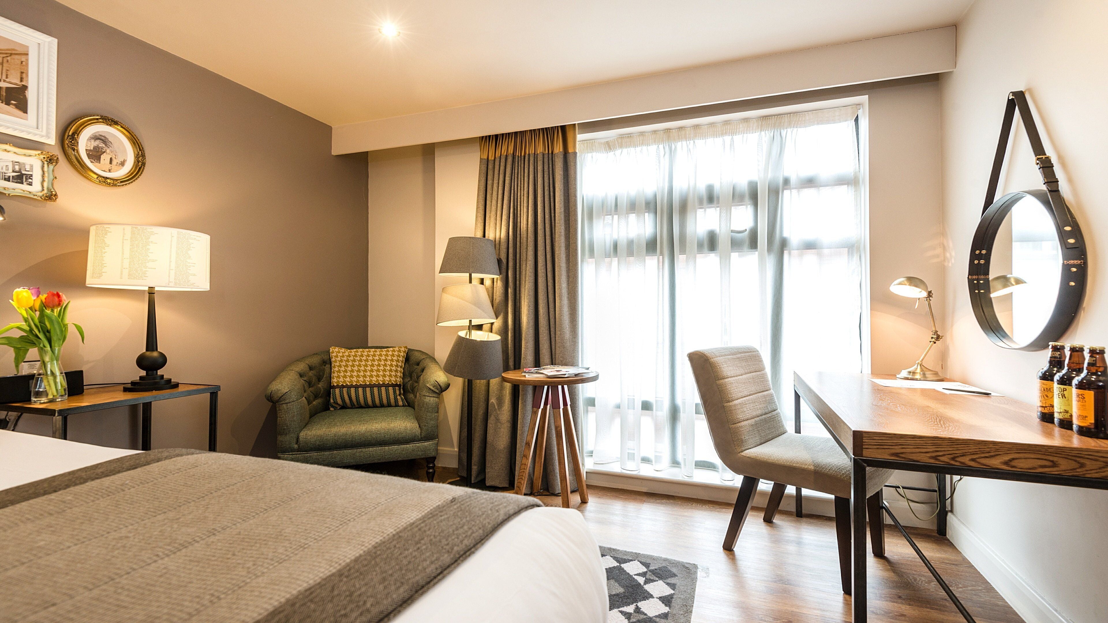 A hotel room with a bed, a desk with a chair, a lamp, a mirror, and a window with curtains.
