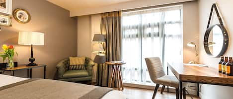 A hotel room with a bed, a desk with a chair, a lamp, a mirror, and a window with curtains.
