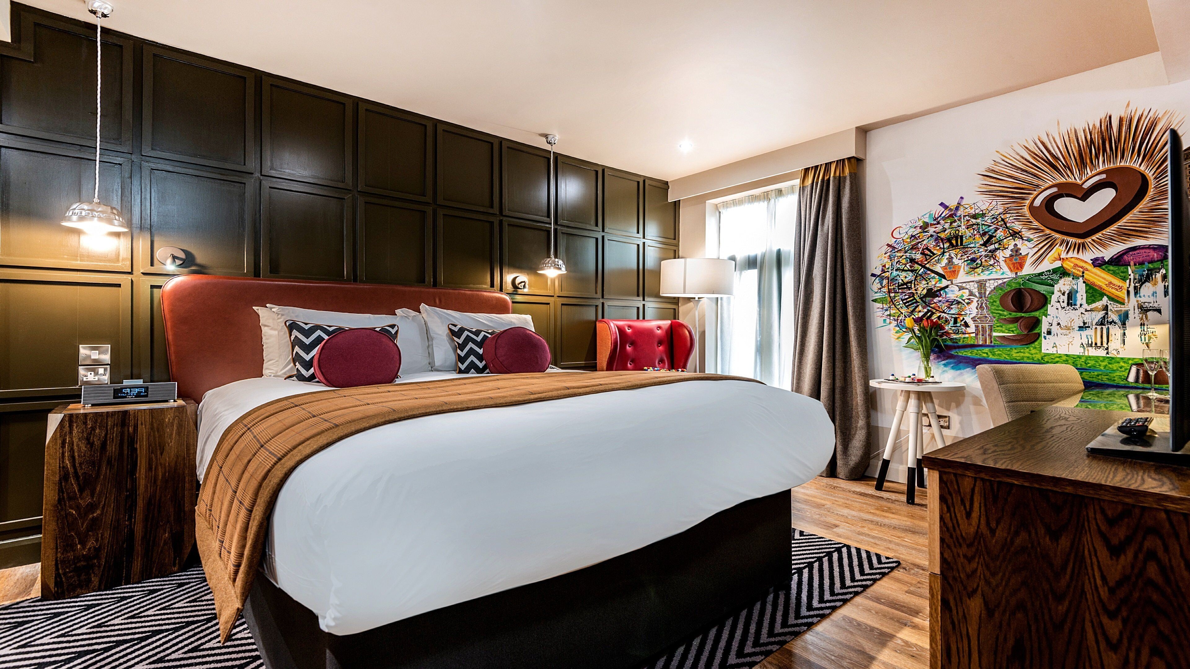 Een hotelkamer met een groot bed, donkerhouten paneelbekleding, een rood beklede hoofdeinde en een kunstwerk met een hartmotief.