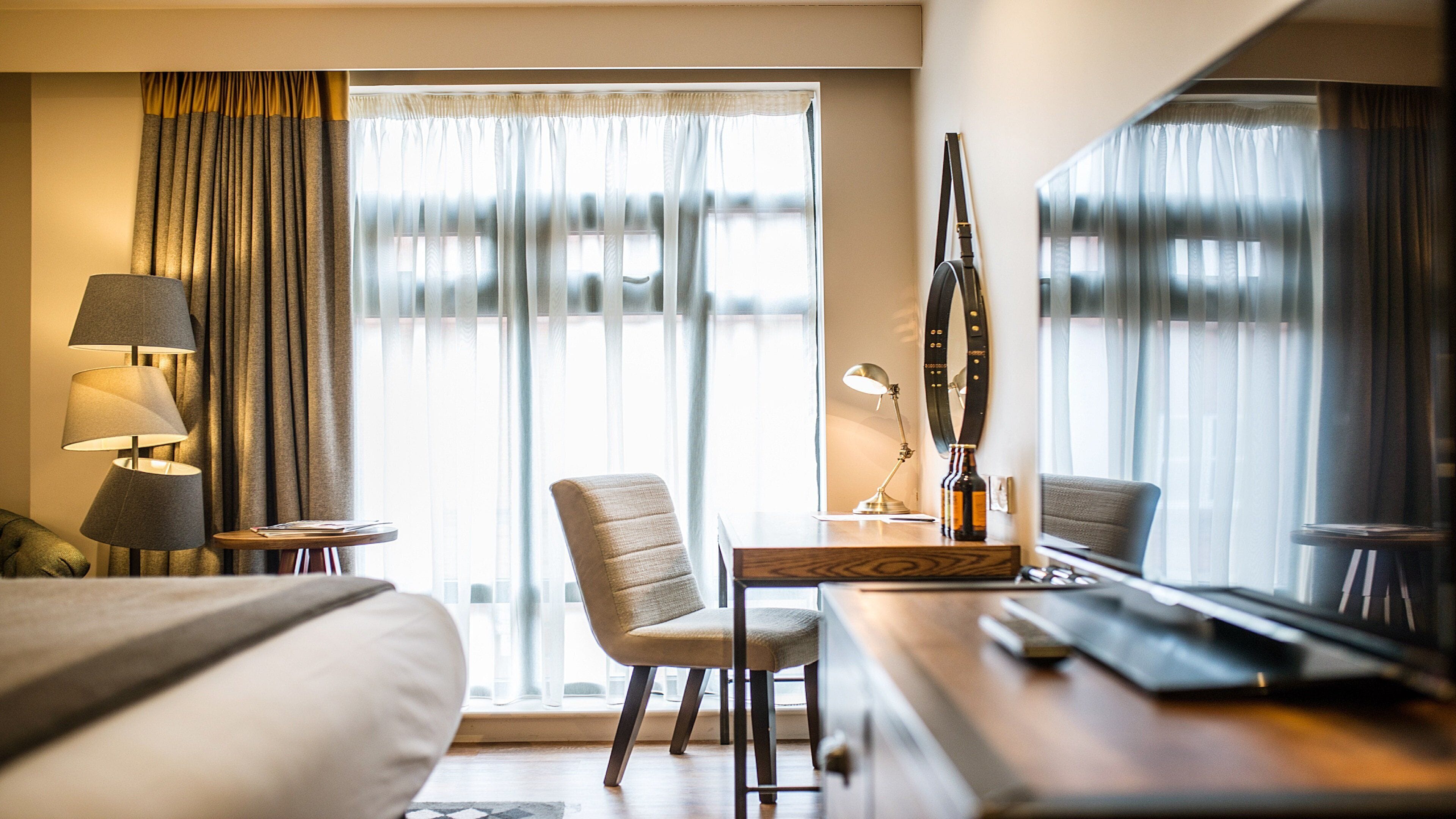 Habitación de hotel con cama, escritorio, silla, lámpara y un amplio ventanal con cortinas.
