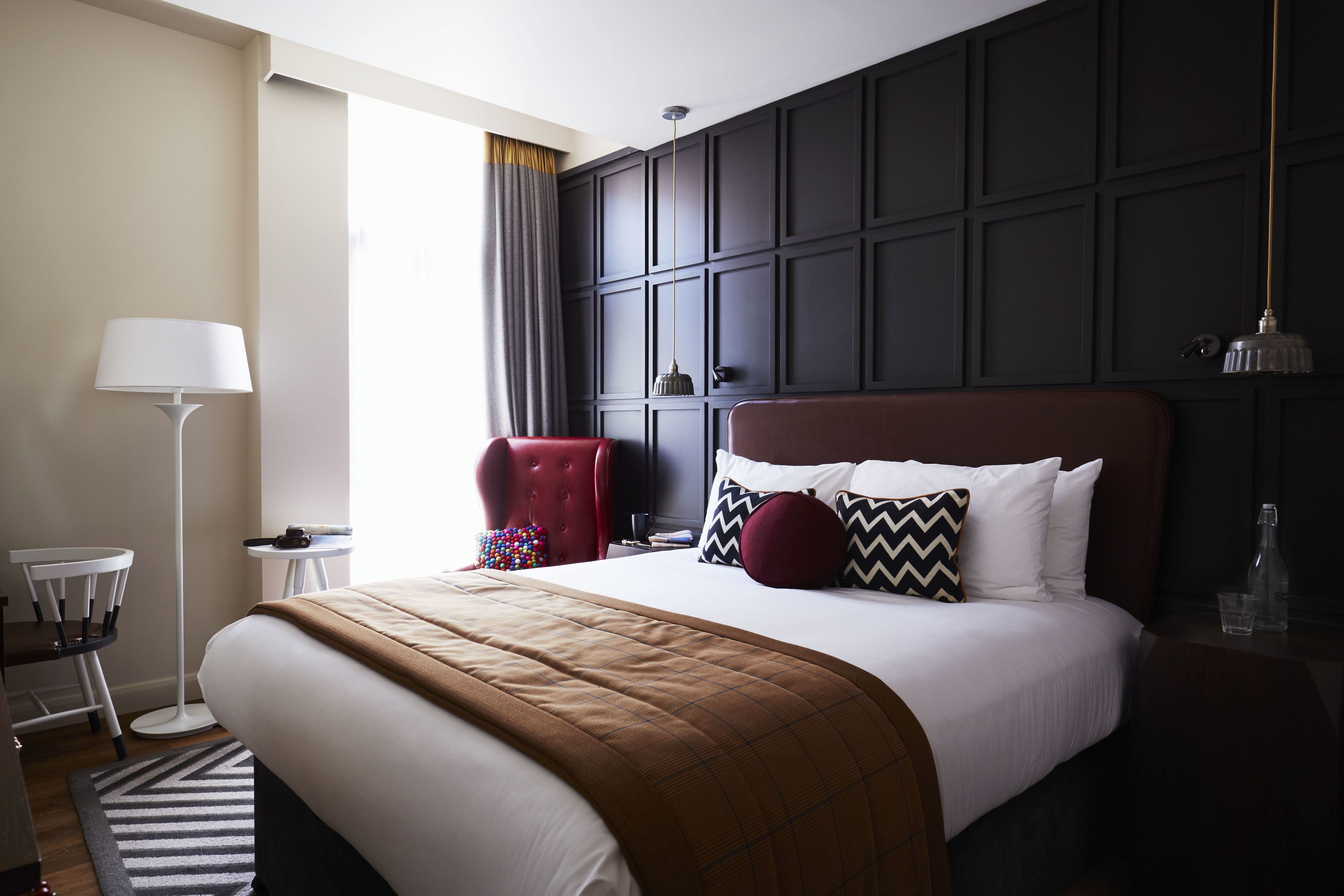 A hotel room with a large bed, a red armchair, a floor lamp, and a view of the outdoors.