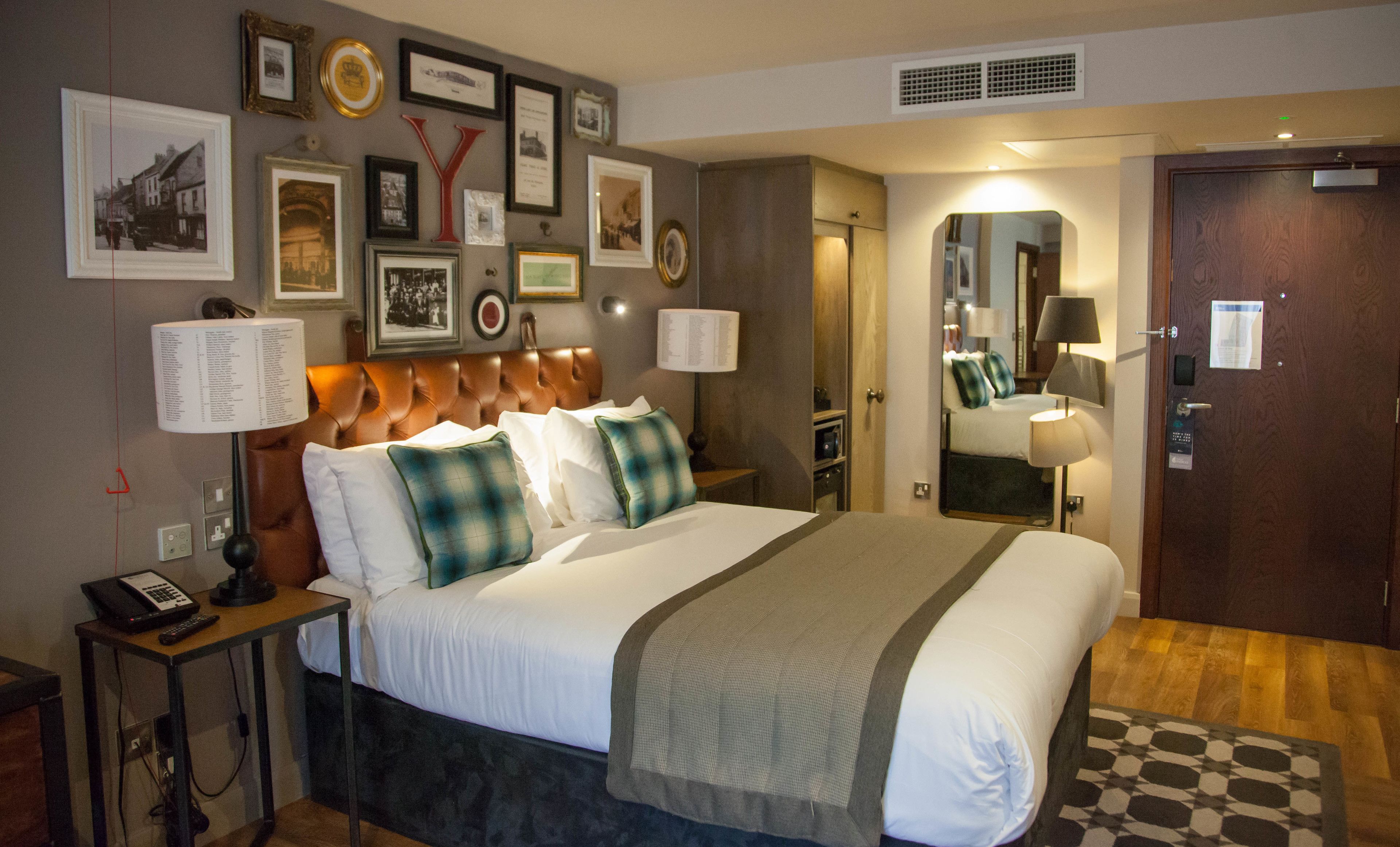 A hotel room with a large bed, a bedside table with a lamp, a mirror, and framed pictures on the wall.