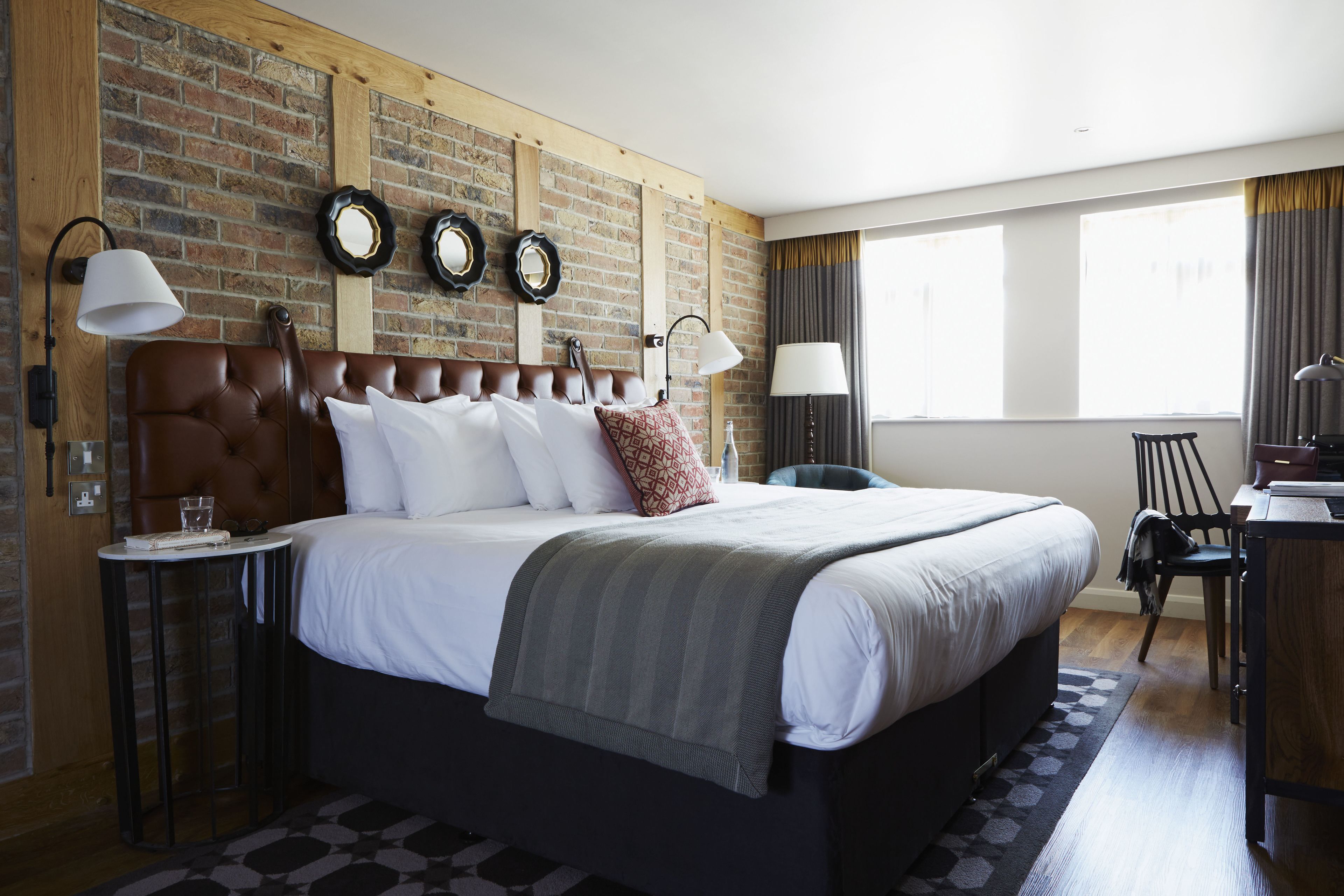 A hotel room with a large bed, two bedside tables, a desk, and a chair. The room features a brick wall, a large window with curtains, and a wooden ceiling.