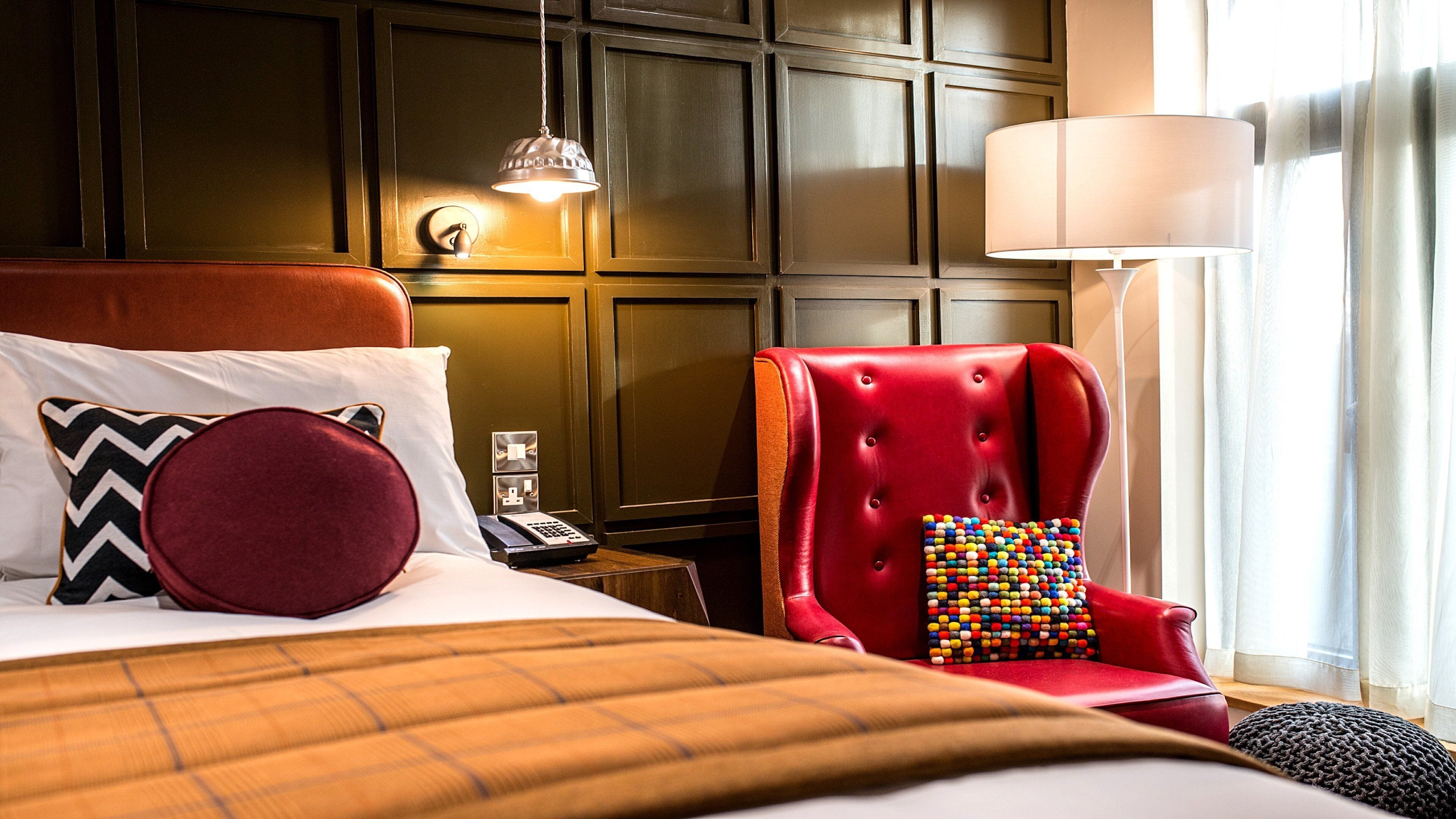 Habitación de hotel con una cama grande, un sillón rojo, una lámpara y vistas al exterior a través de una ventana.