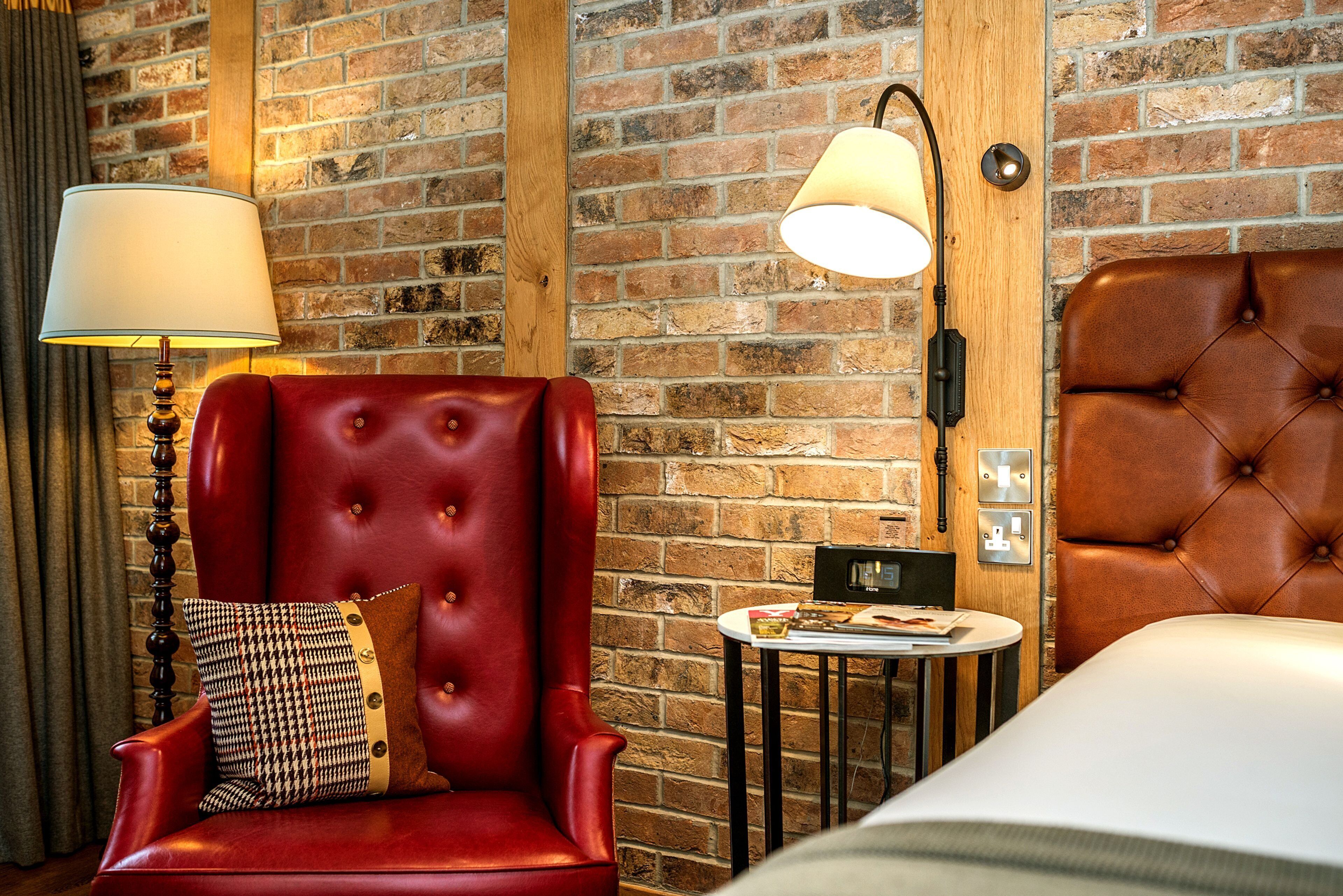 A hotel room with a brick wall, a red leather armchair, a white bed, a small round table, and a lamp.