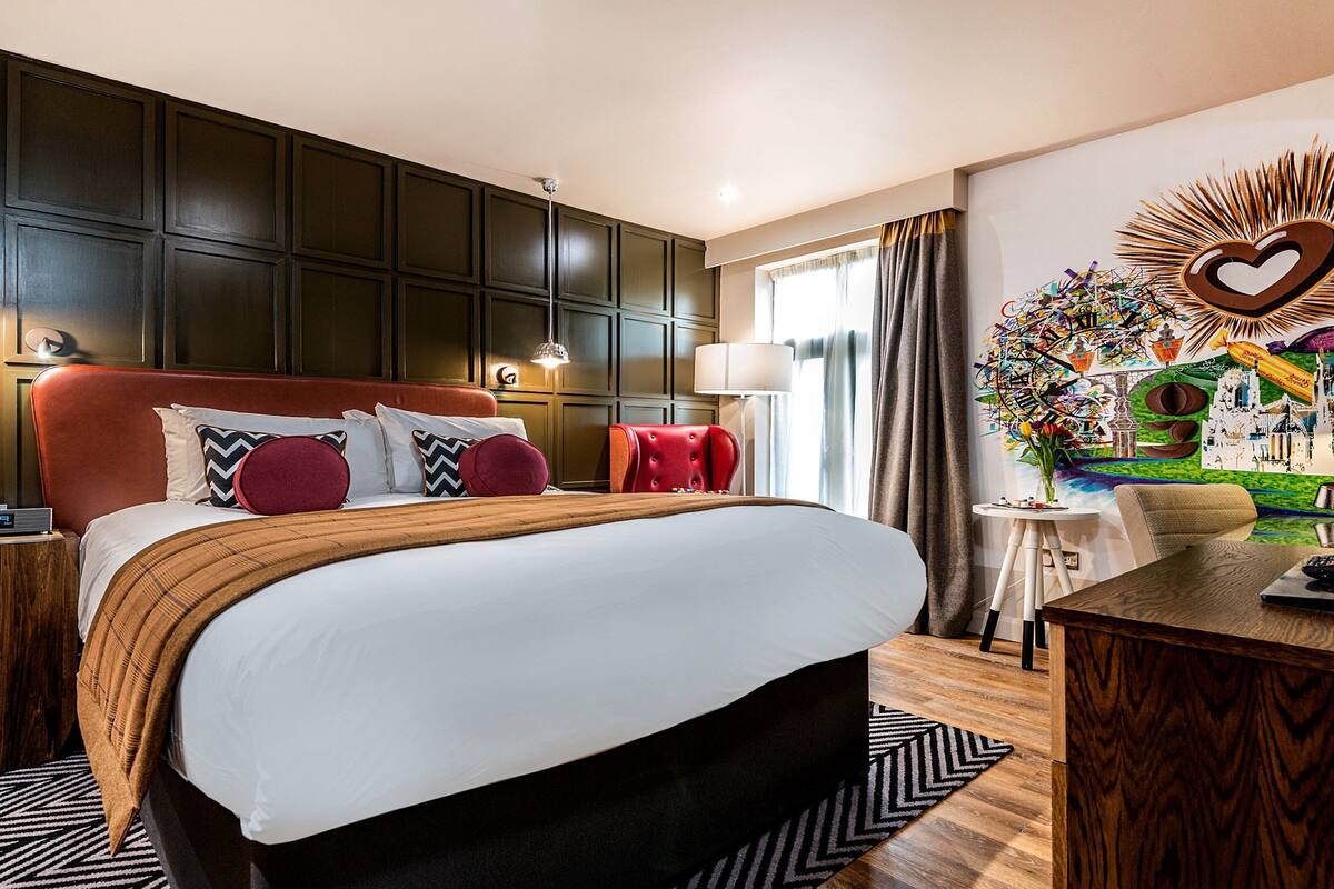 A hotel room with a large bed, dark wooden paneling, a red upholstered headboard, and a wall art piece with a heart design.