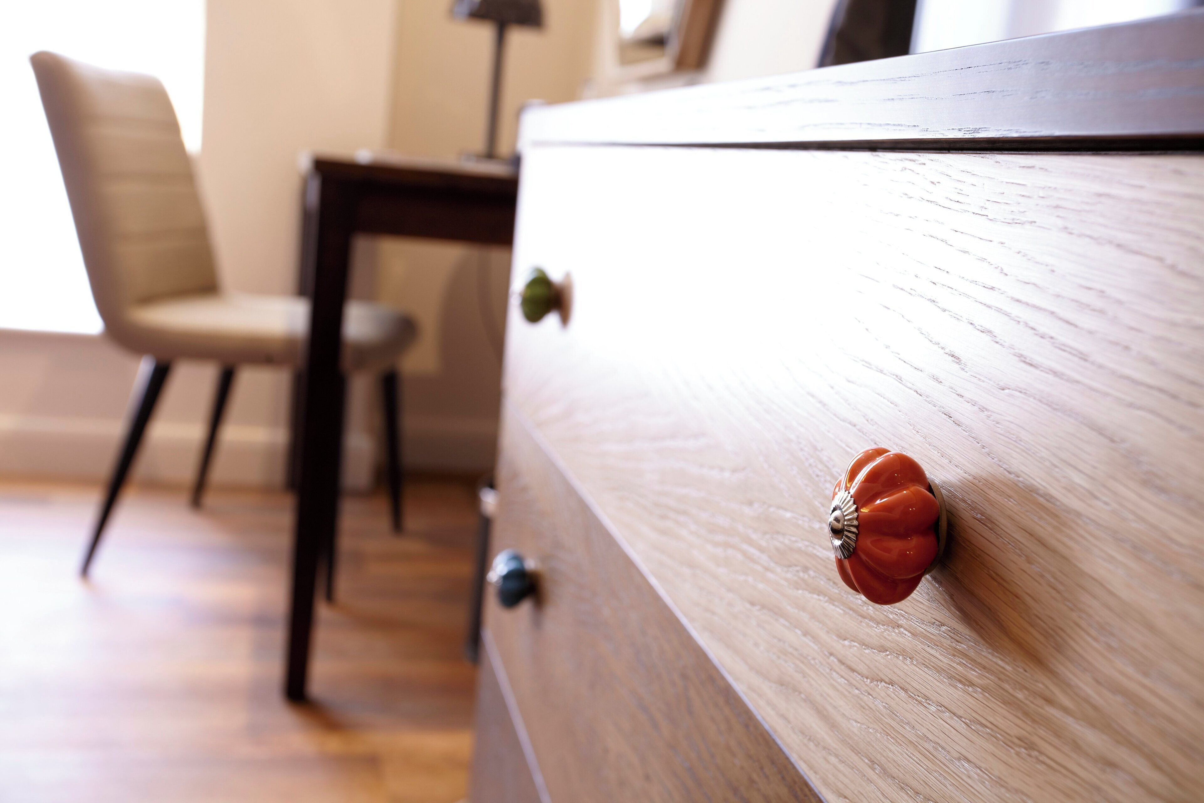 A wooden dresser with a floral knob, a chair, and a lamp in a room with wooden flooring.