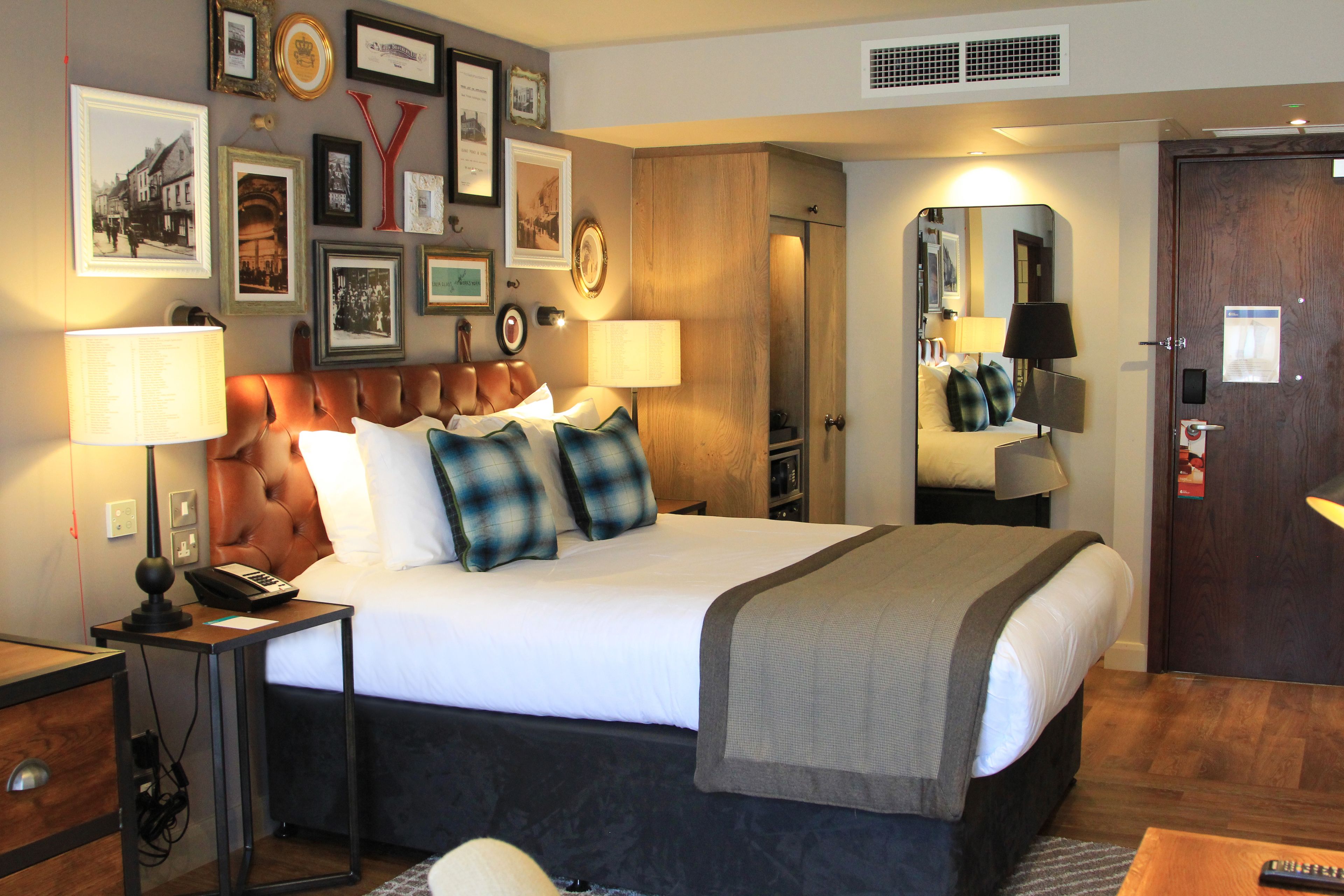 Habitación de hotel con una cama grande, lámparas de noche, cabecero de madera, un espejo y un muro decorado con cuadros.