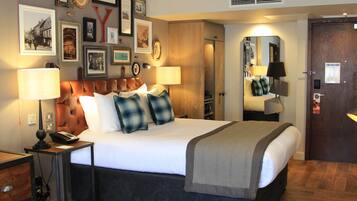 Habitación de hotel con una cama grande, lámparas de noche, cabecero de madera, un espejo y un muro decorado con cuadros.