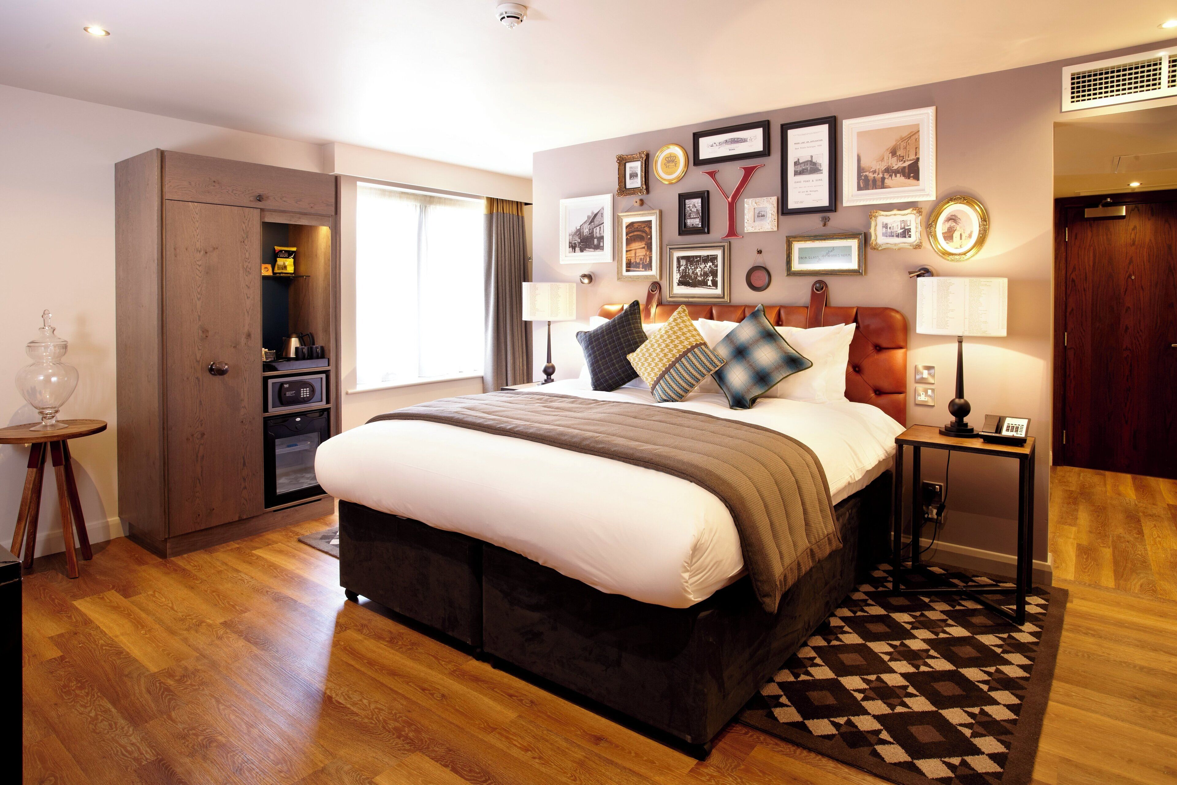 A hotel room with a large bed, a wooden wardrobe, a small table with a lamp, and a wall decorated with framed pictures.