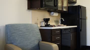 A compact kitchen with a microwave, stove, and refrigerator. There is a chair with a blue cushion and a dishwasher.