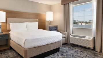 A hotel room with a large bed, a nightstand, a chair, a window with curtains, and a wall-mounted air conditioner.