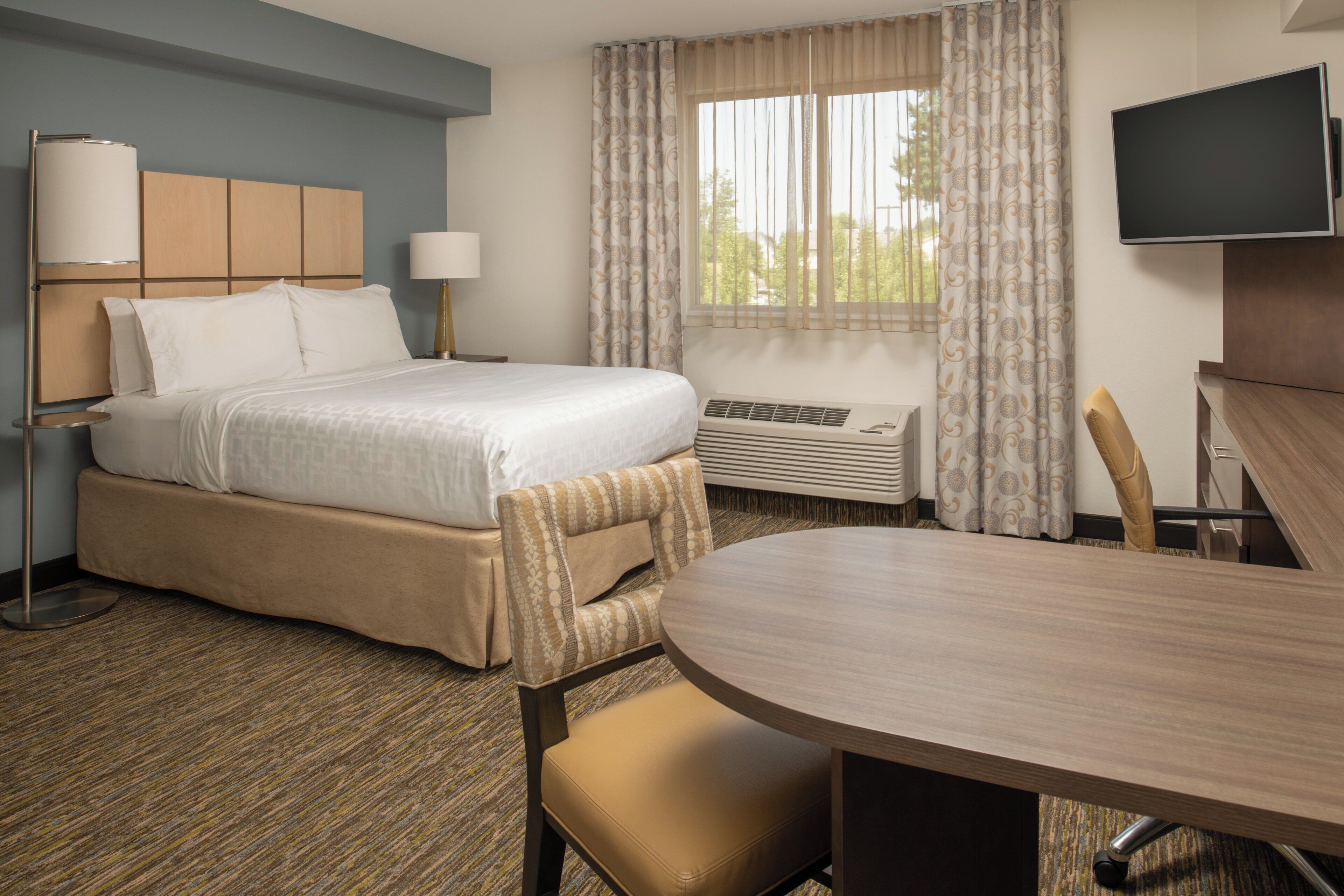 A hotel room with a bed, bedside table, lamp, air conditioning unit, television, and a dining table with chairs.
