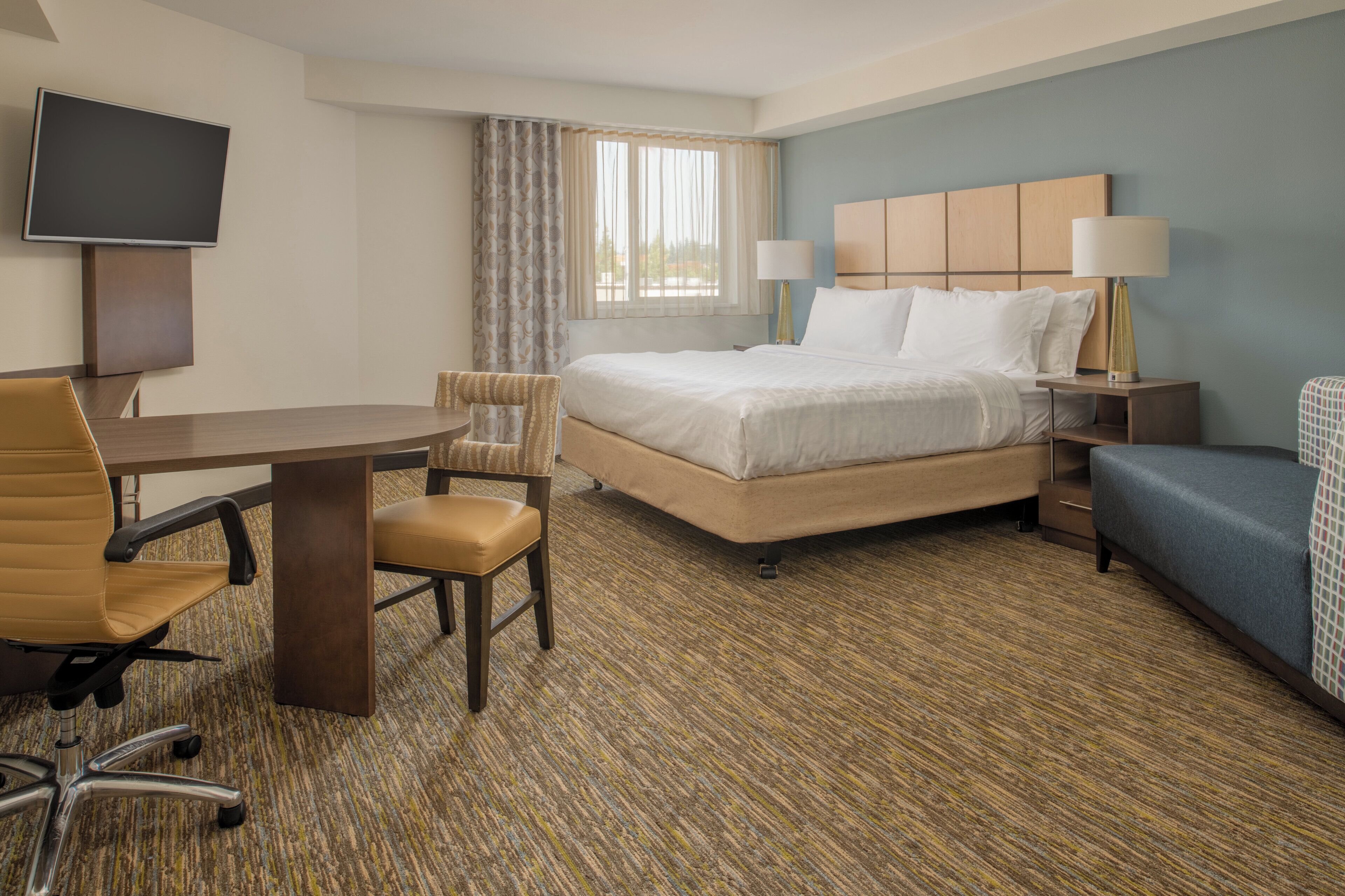 Una habitación de hotel con cama, escritorio, silla, televisor y ventana con cortinas.