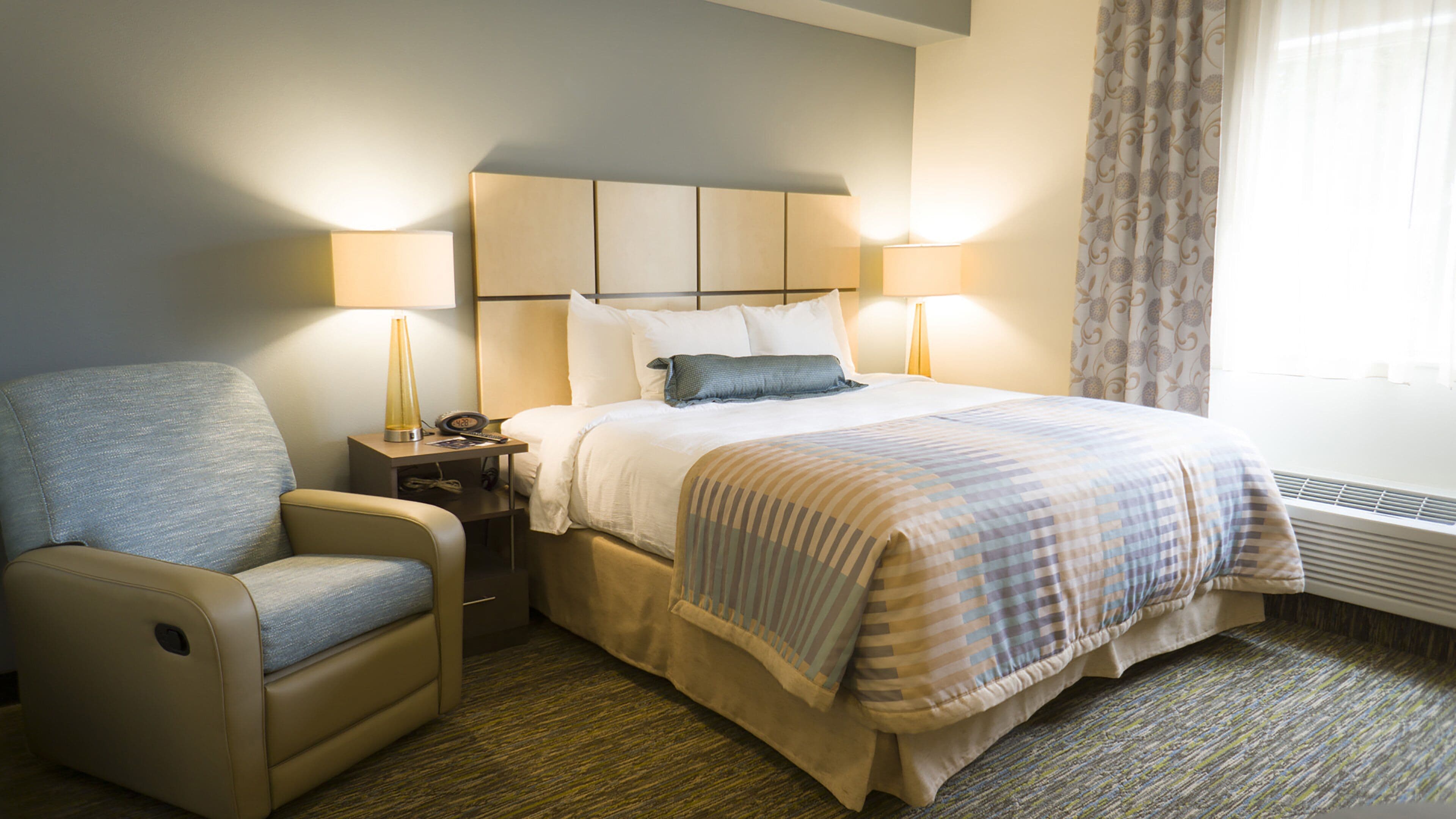 A hotel room with a bed, a chair, a nightstand, and a lamp.
