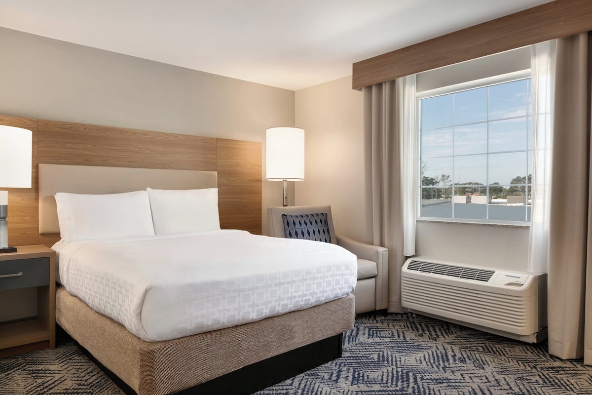 Una habitación de hotel con una cama grande, una mesita de noche, una silla, una ventana con cortinas y un aire acondicionado instalado en la pared.